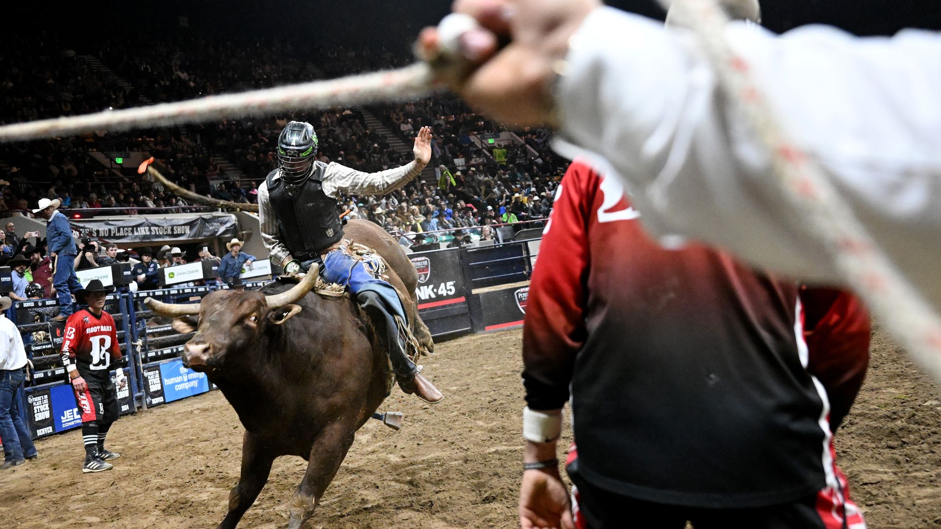 A man wearing a vest and helmet rides a bucking bull while a man holds a rope in the foreground. 
