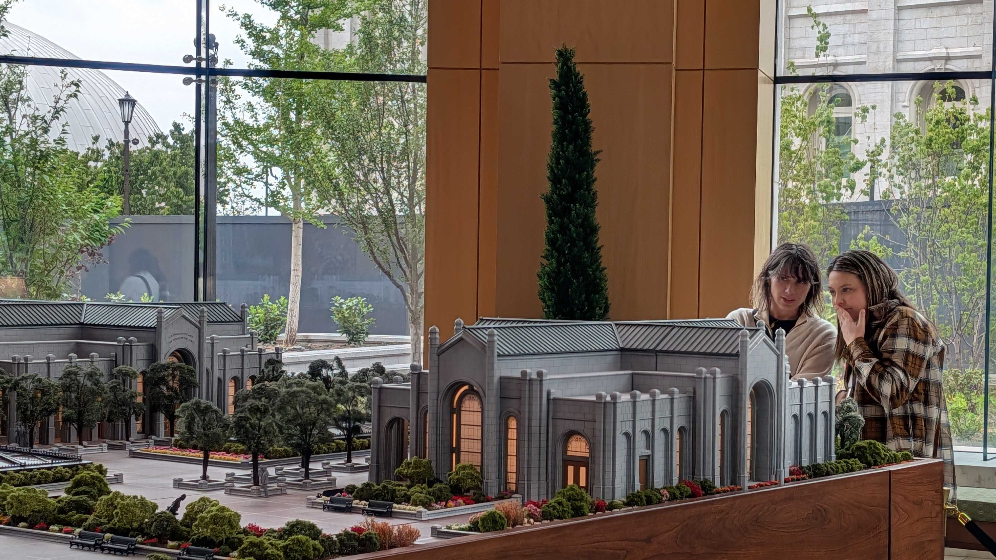 Indoor scene showing a detailed miniature gray stone building complex with arches, courtyards, and manicured trees. Two women in plaid jackets study the model; glass windows reveal greenery outside.