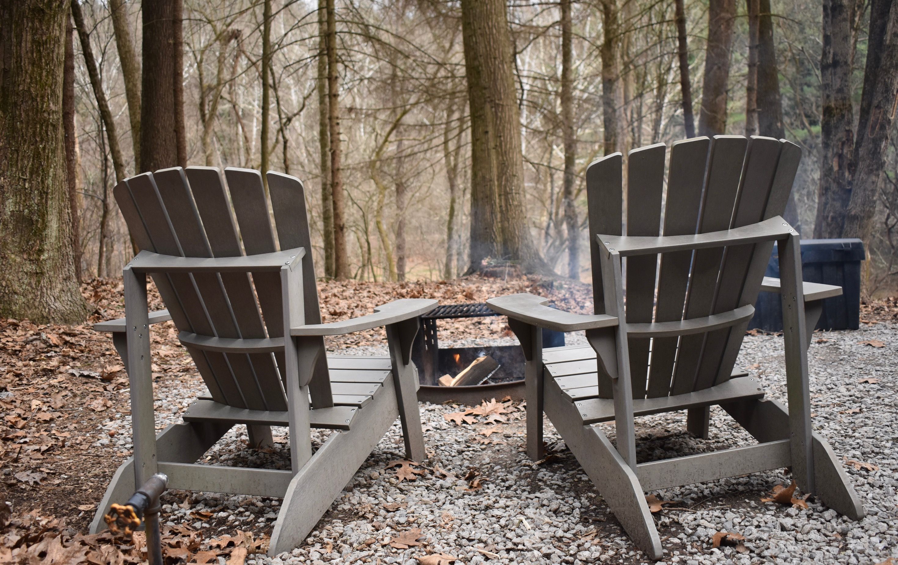 Two gray Adirondack chairs flank a small fire pit on a gravel clearing, surrounded by a leaf-strewn forest as smoke rises and trees loom in the background.