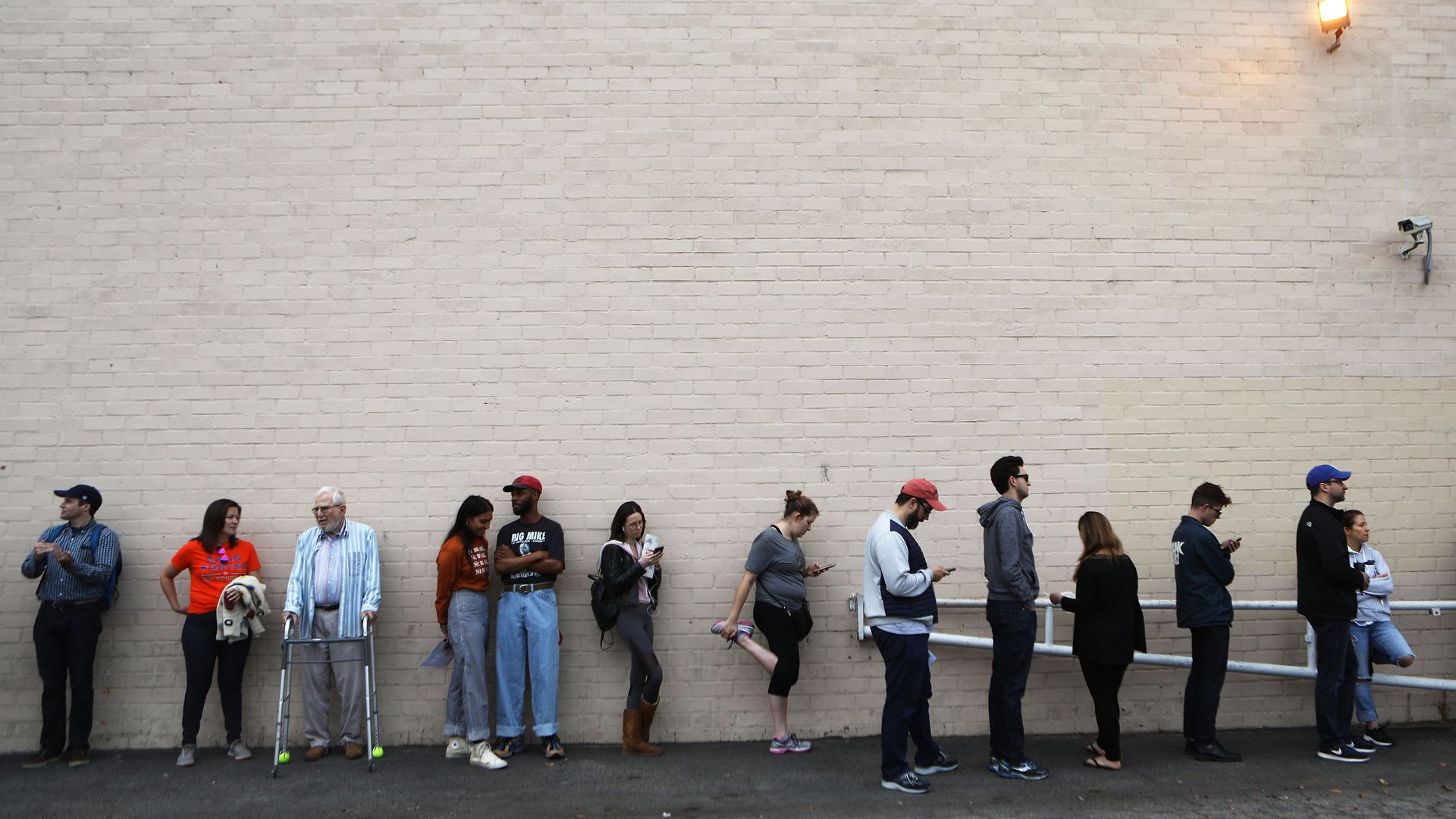 People stand in line to vote at a polling site. There is an old man with a walker and one woman stretching her legs.