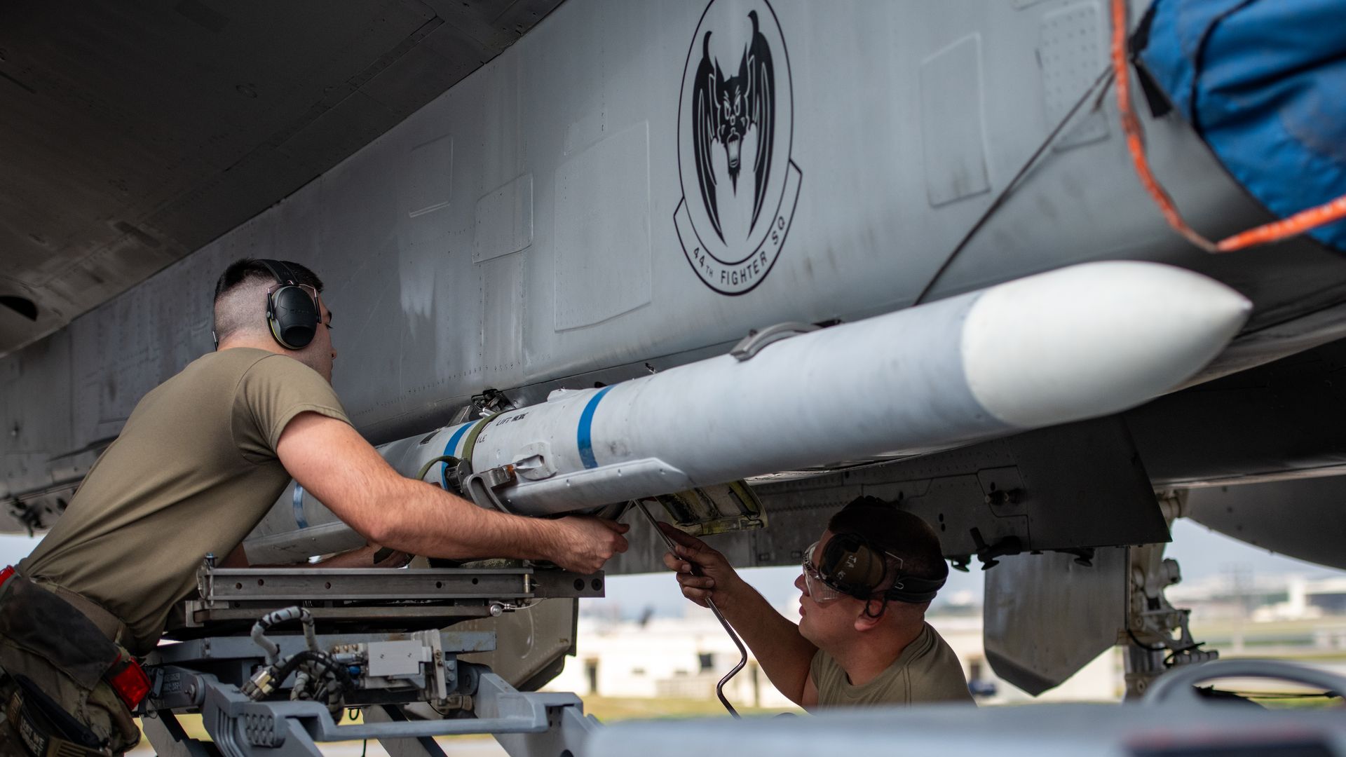 Two military personnel in tan shirts and hearing protection work on attaching a white missile to a gray jet with a black bat emblem of the 49th Fighter Sq on its side.