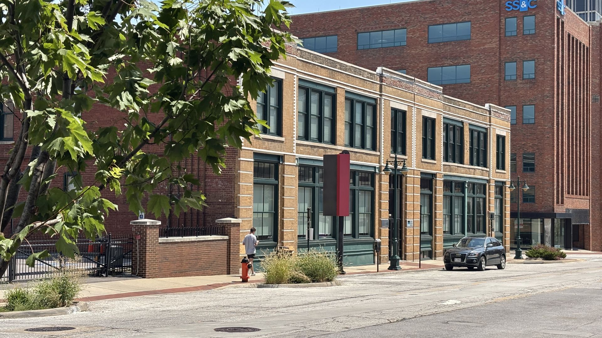 Tree in the foreground partially hides a brick street with storefronts and green window frames. A man stands near a red hydrant; a car is parked. A tall brick building with SS&C rises in the background.
