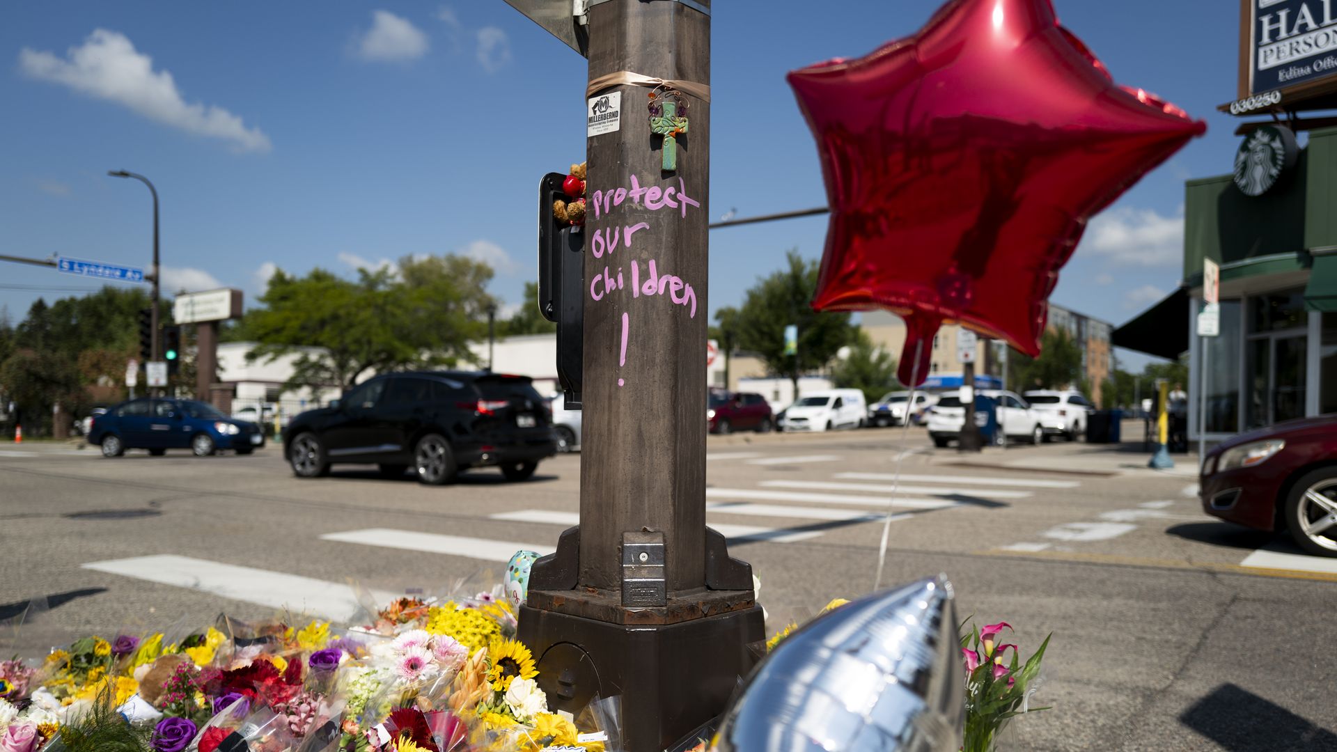 Memorial at street corner with flowers, red star-shaped and silver balloons tied to a pole with pink chalk saying "Protect our children!", cars and blue sky in background.