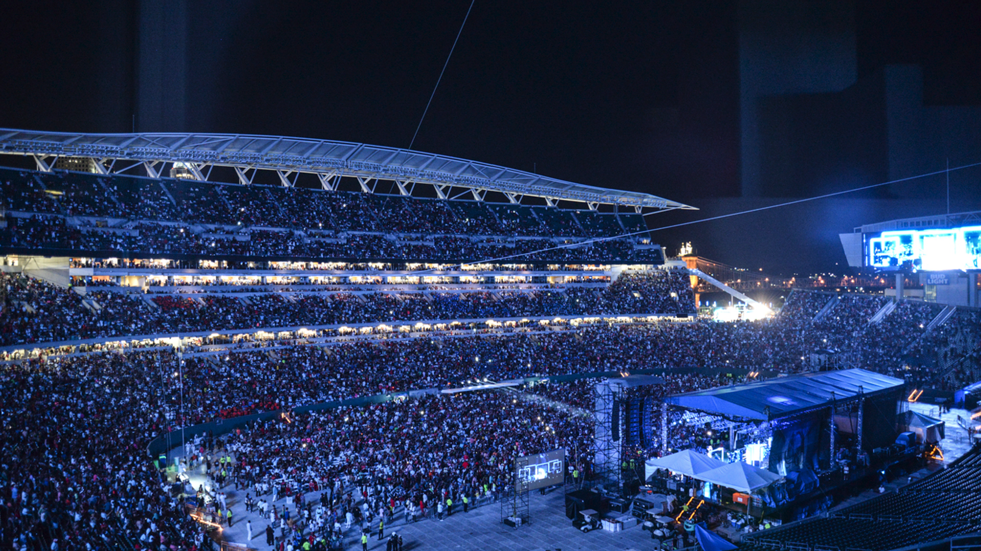 Nighttime stadium filled with a massive crowd across multiple tiers. A brightly lit stage and blue-screen displays dominate the right; tents and equipment line the front, dark sky overhead.