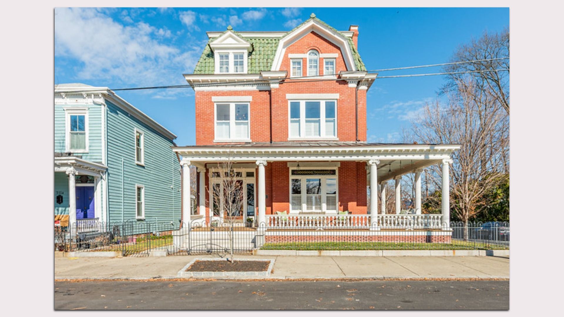 Large red brick house with green shingled roof, white columns, and wrap-around porch in a suburban neighborhood under a blue sky.