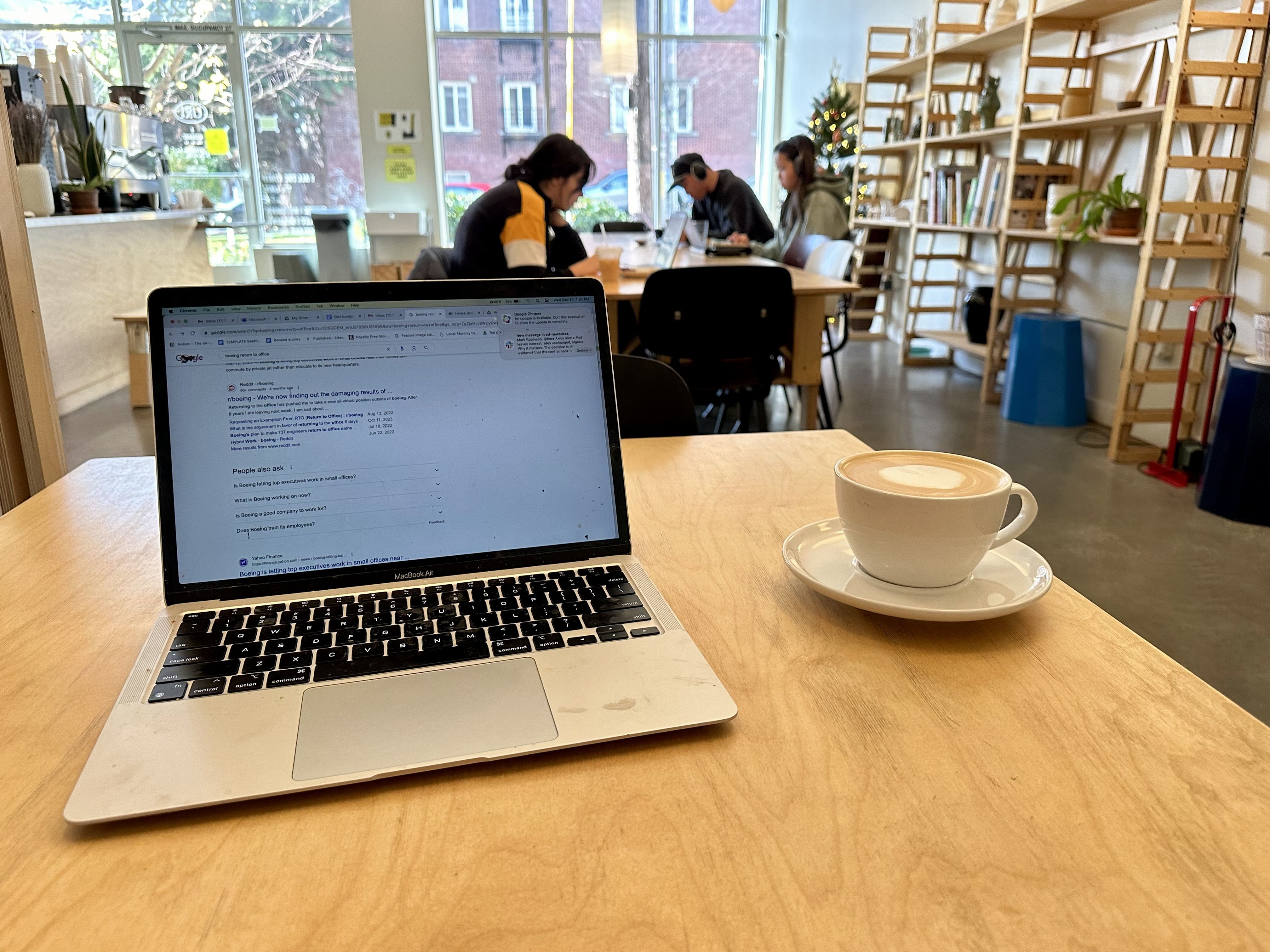 A laptop and a cappuccino sitting on a light colored wood table with bookshelves and people working in background.