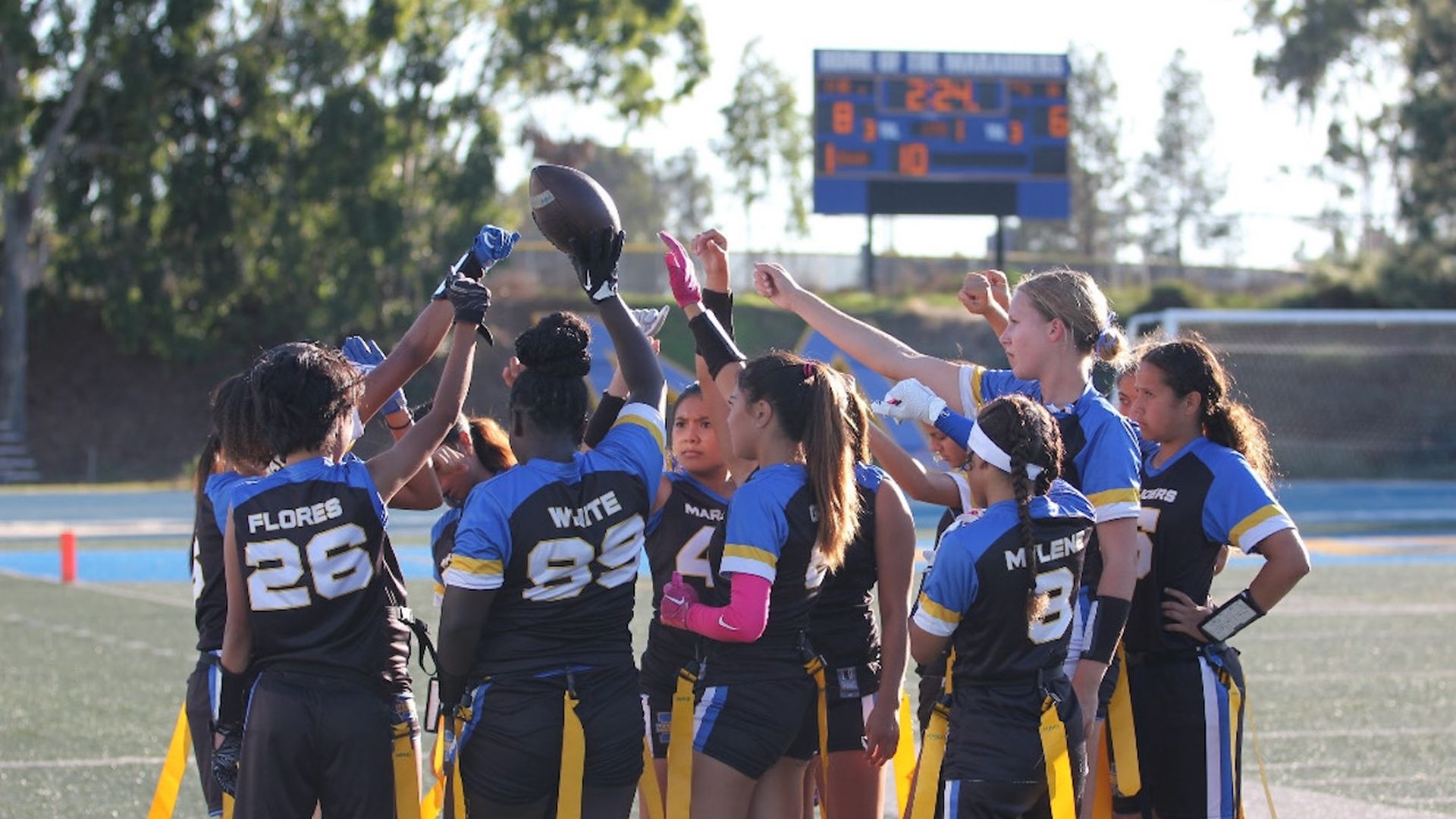 Girls' flag football team huddles on a turf field, raising hands in celebration. They wear blue and black uniforms with yellow sashes; a scoreboard and trees are in the background.