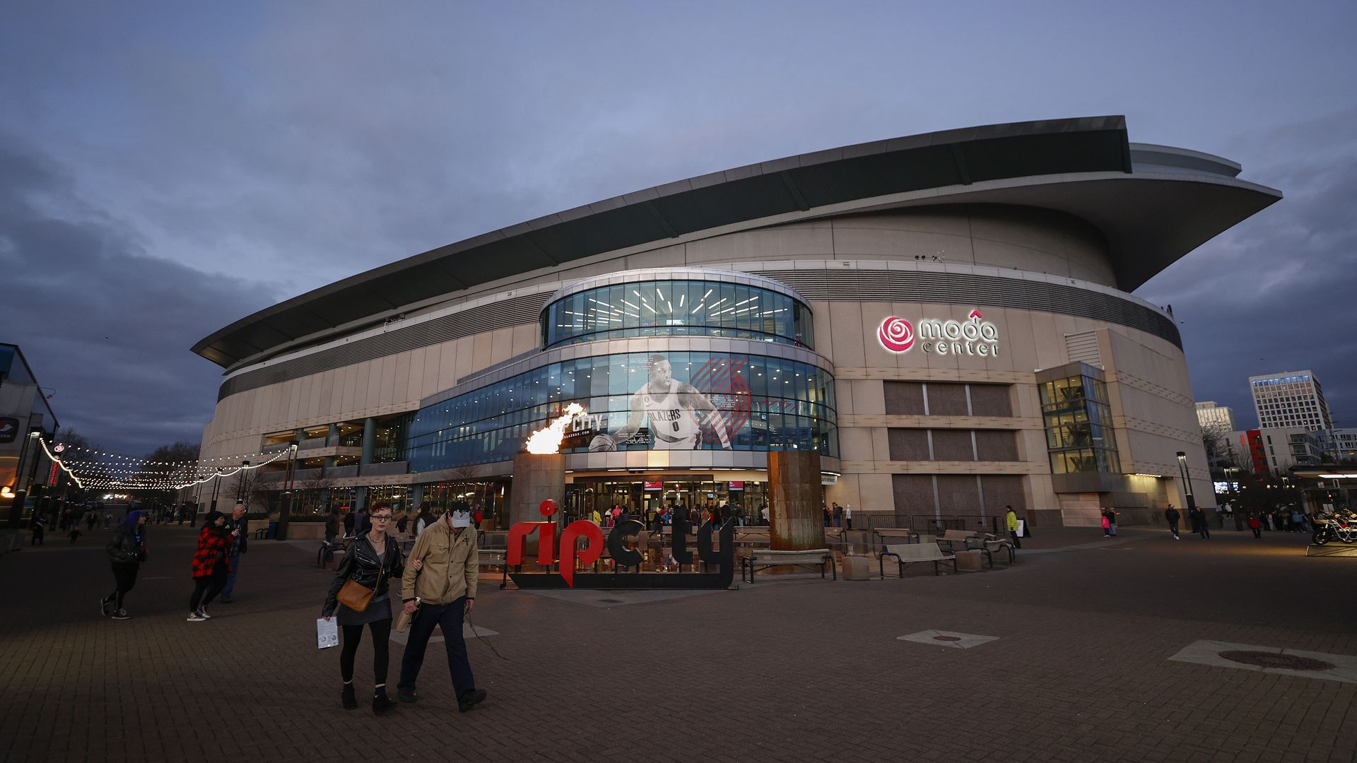 Portland's rounded arena the Moda Center with a black and red Rip City sign in front and two people walking past.