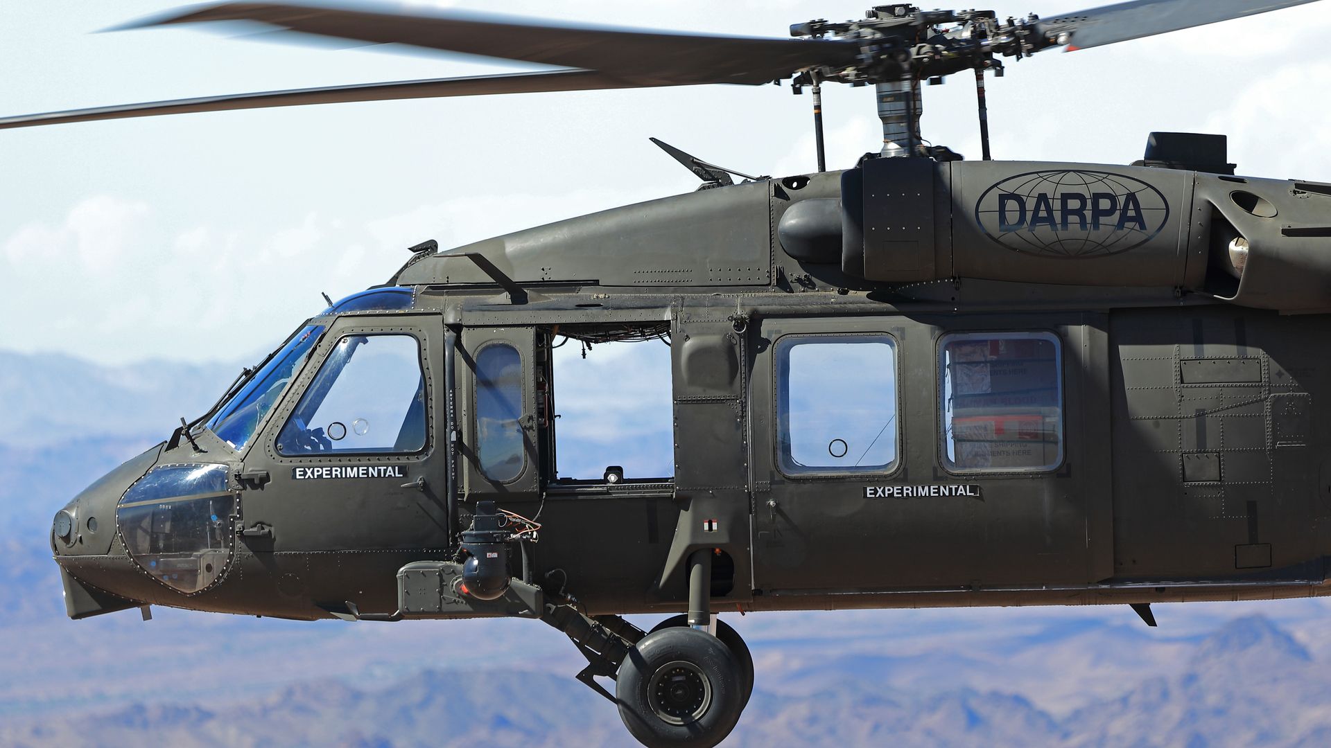 A Black Hawk helicopter, flying without a pilot. Mountains are seen in the back. The DARPA logo is stamped on the side of the helicopter.