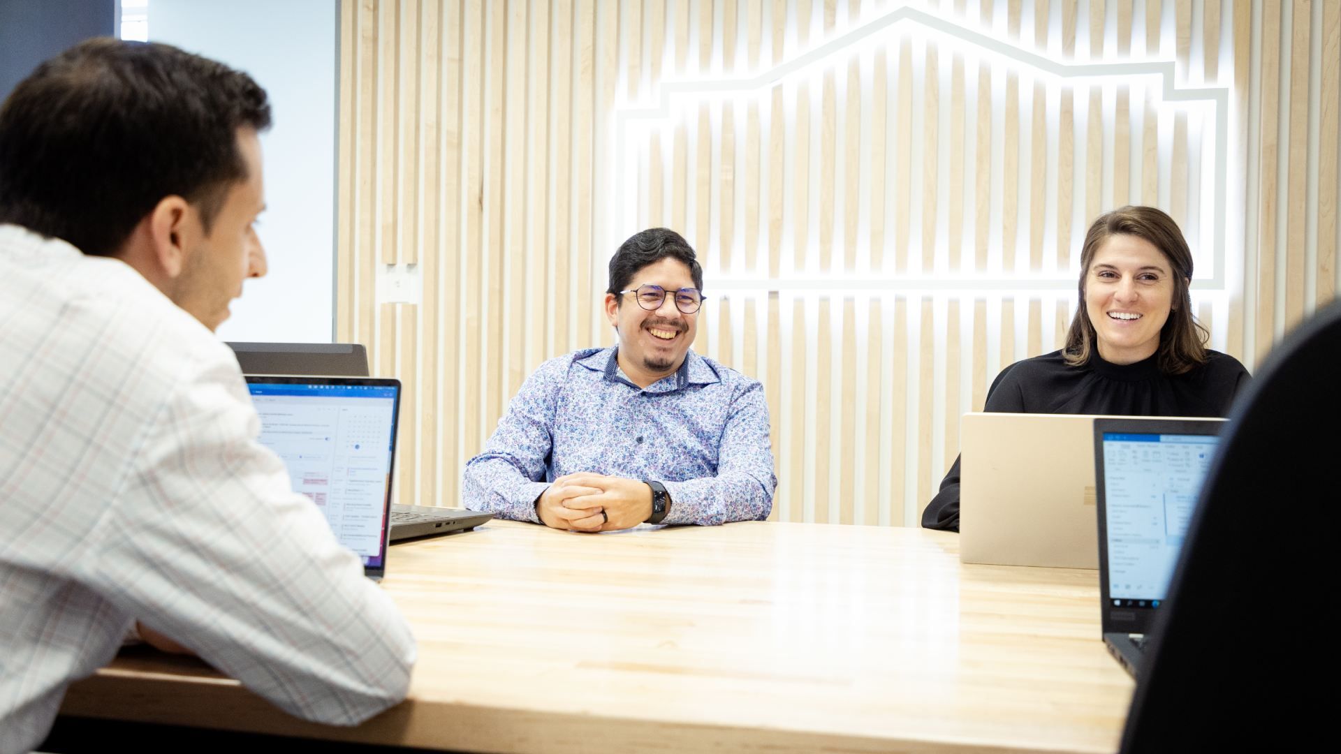 Three coworkers sit around a light wood table in a bright office, smiling and talking. Center is a man with glasses in a blue patterned shirt; to his right a woman in black; laptops open.