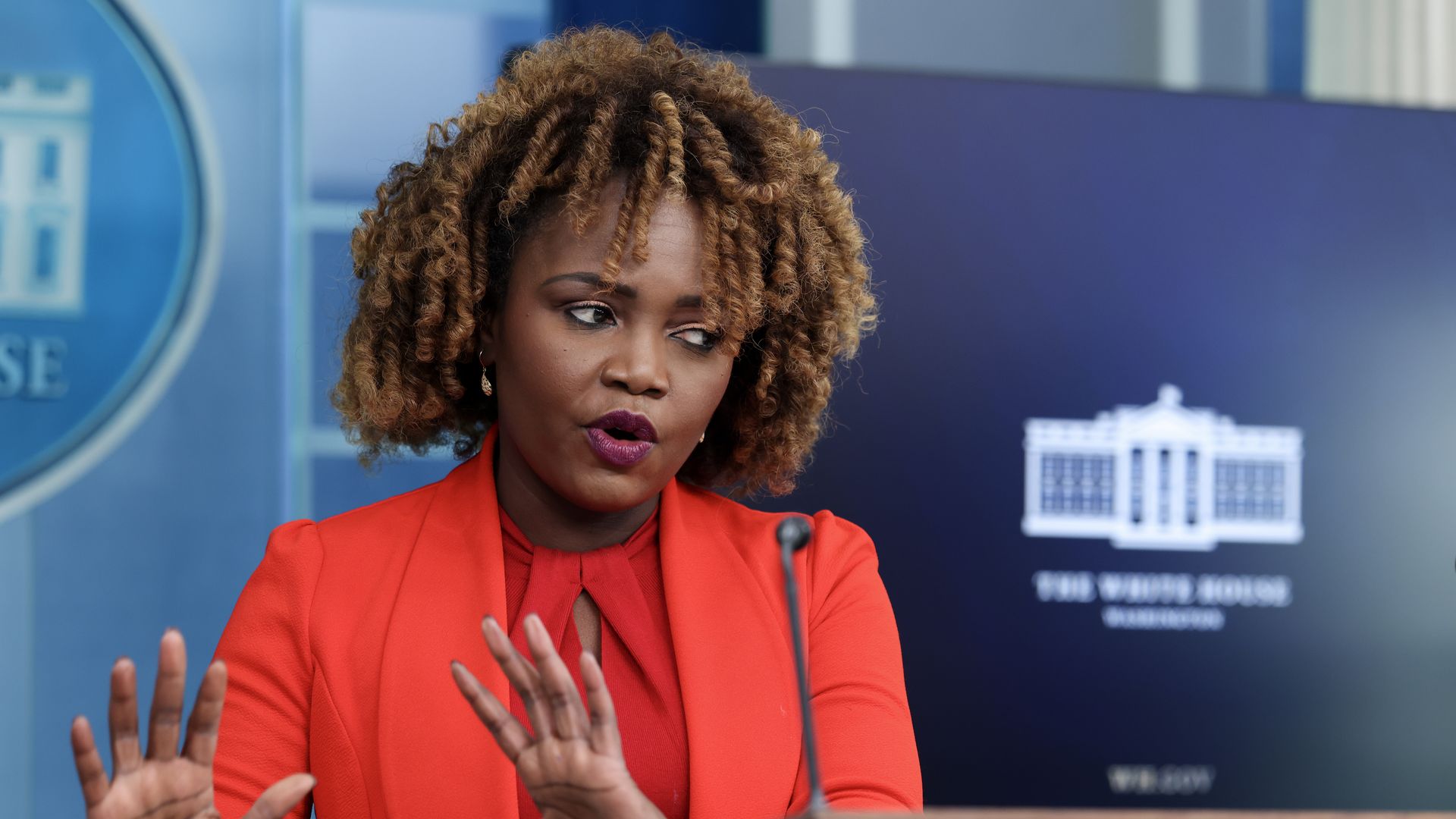 Karine Jean Pierre, wearing a red blazer and standing in front of a screen with the White House logo on it.