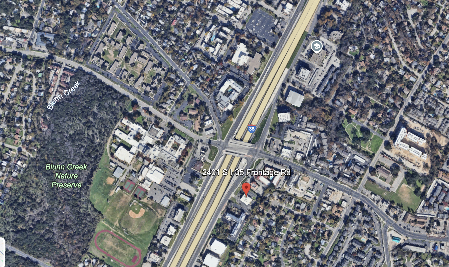 Aerial view of 2401 S I-35 Frontage Rd near Blunn Creek Nature Preserve, showing highway I-35, residential and commercial buildings, green park spaces, and a running track.
