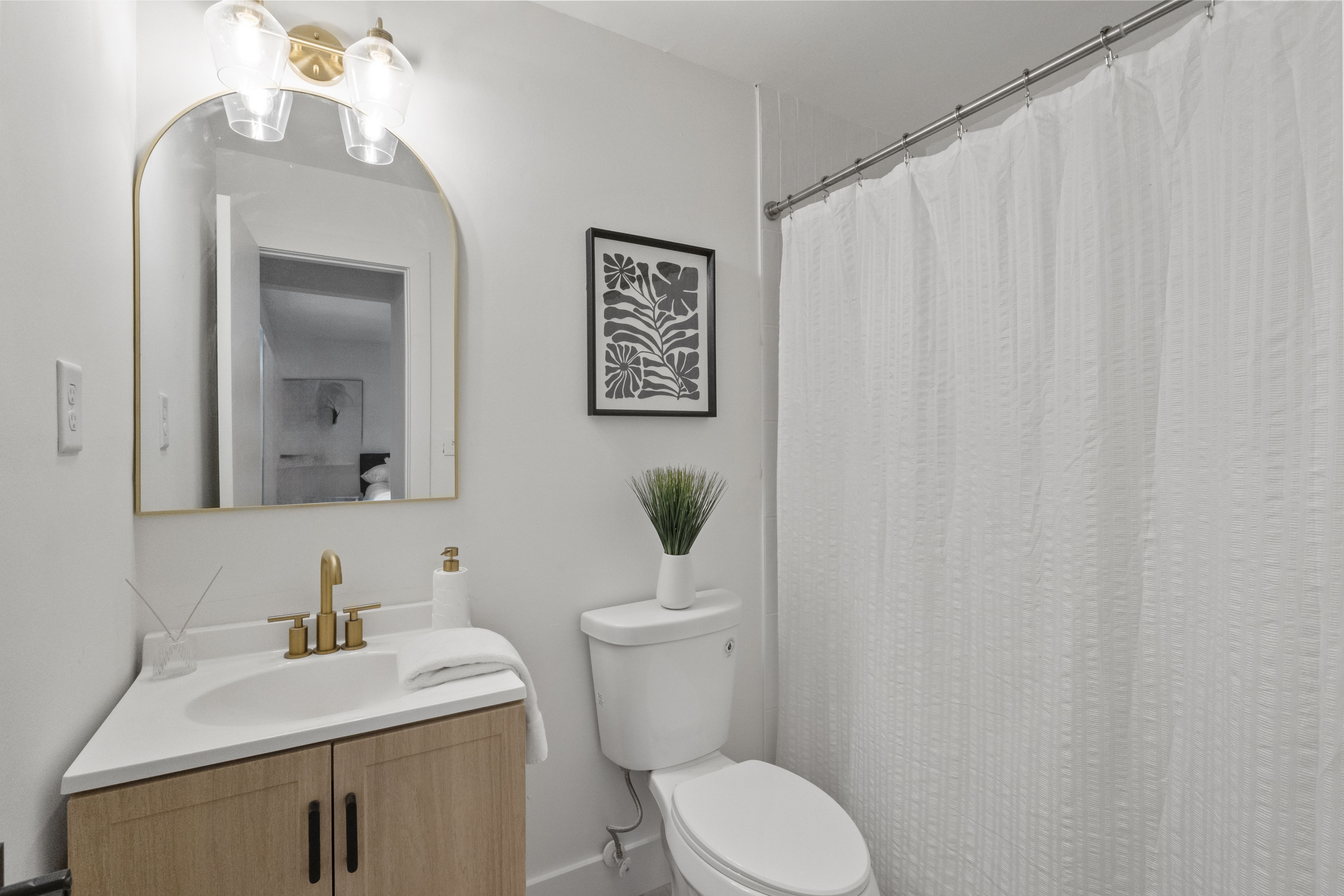 Small modern bathroom with white walls, wooden vanity with brass faucet, arched mirror, white toilet with potted green plant, black and white framed leaf art, and white textured shower curtain.