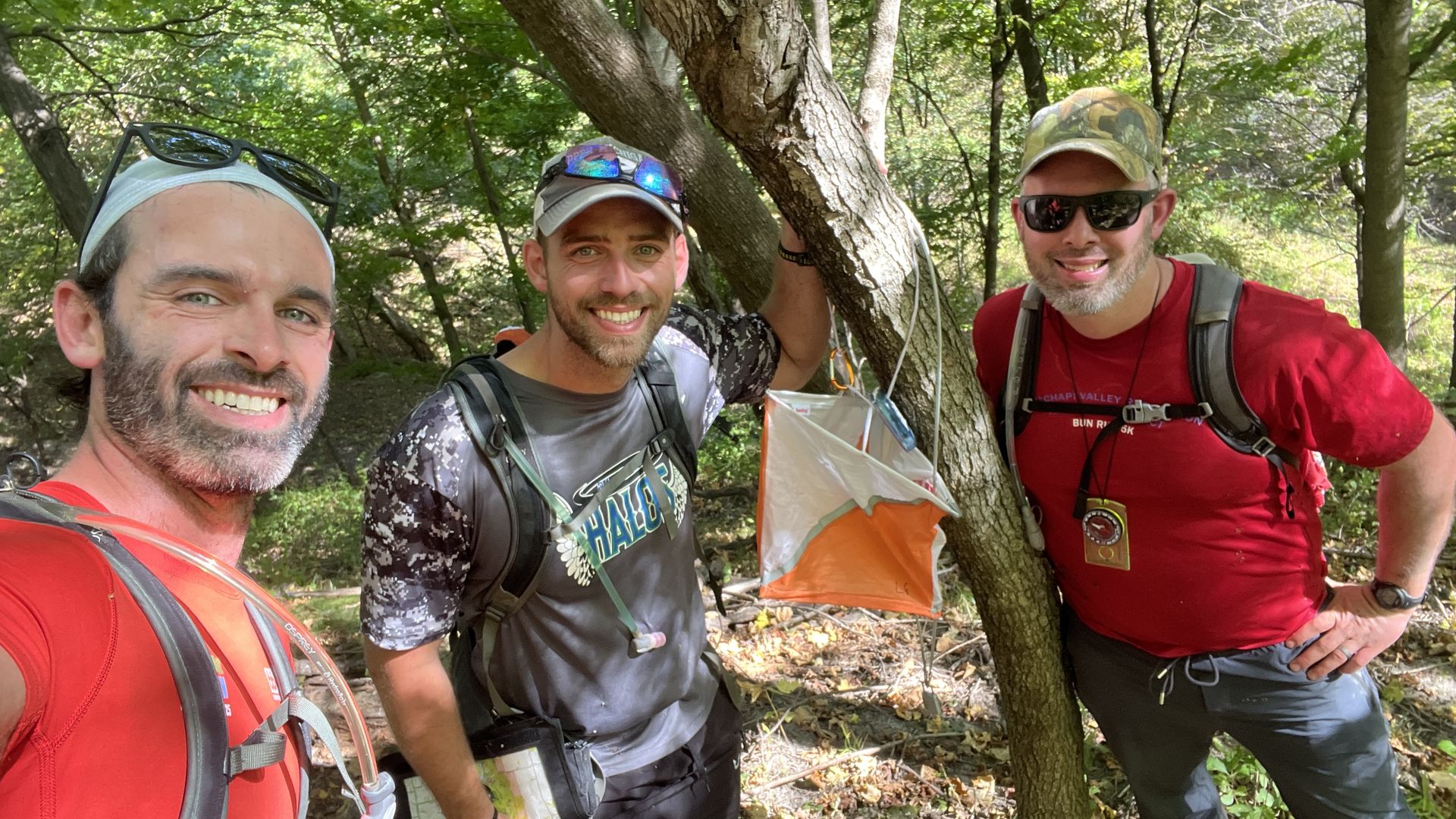 Reporter Travis with his friend and brother smiling in outdoor gear standing in a wooded area next to an orange and white orienteering marker hanging from a tree during the Wild Driftless Adventure Race in Iowa.