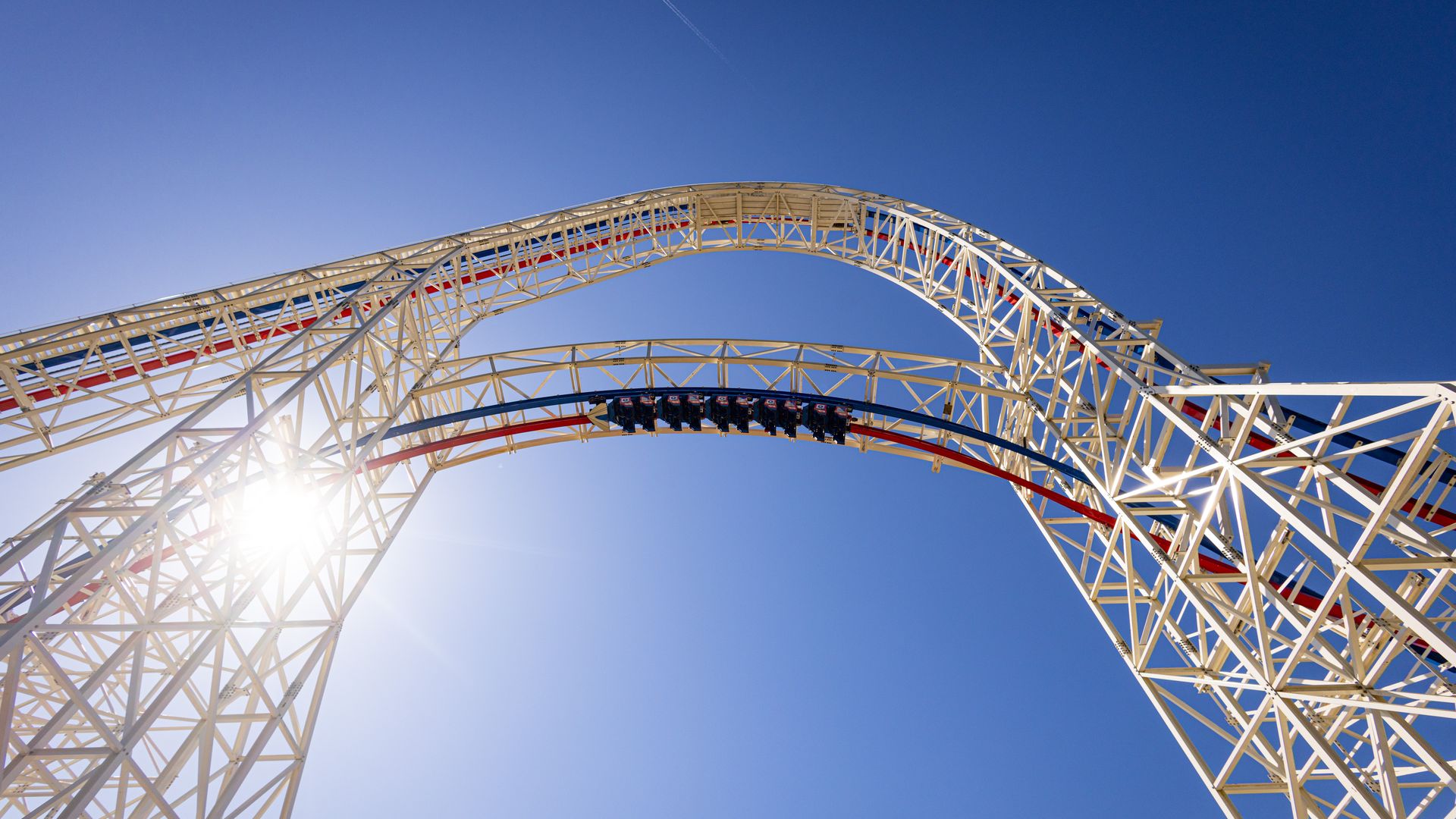 Low-angle shot of a white steel roller coaster track arching across a bright blue sky, with red rails and lattice supports; a train runs along the lower arc as sun glints.