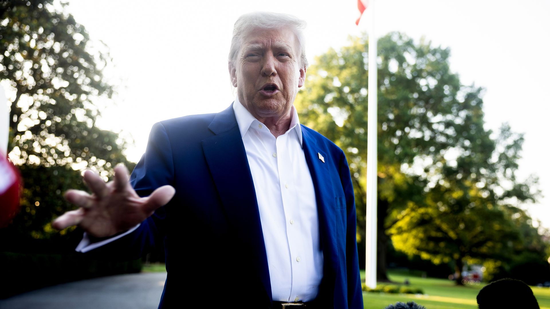 Donald Trump wearing a blue tieless suit in front of trees and a flagpole while speaking into microphnes.