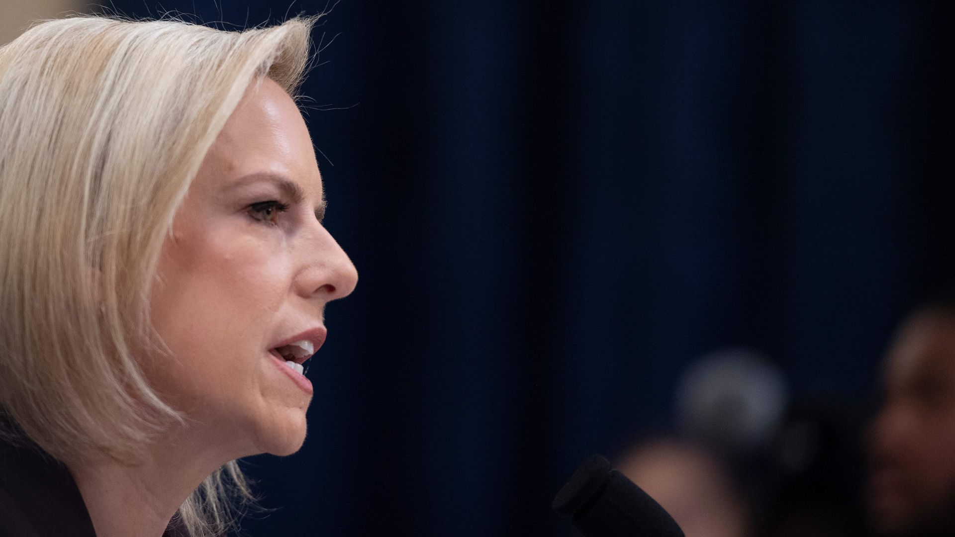 DHS Secretary Kirstjen Nielsen's profile as she speaks into a microphone with a serious face. 