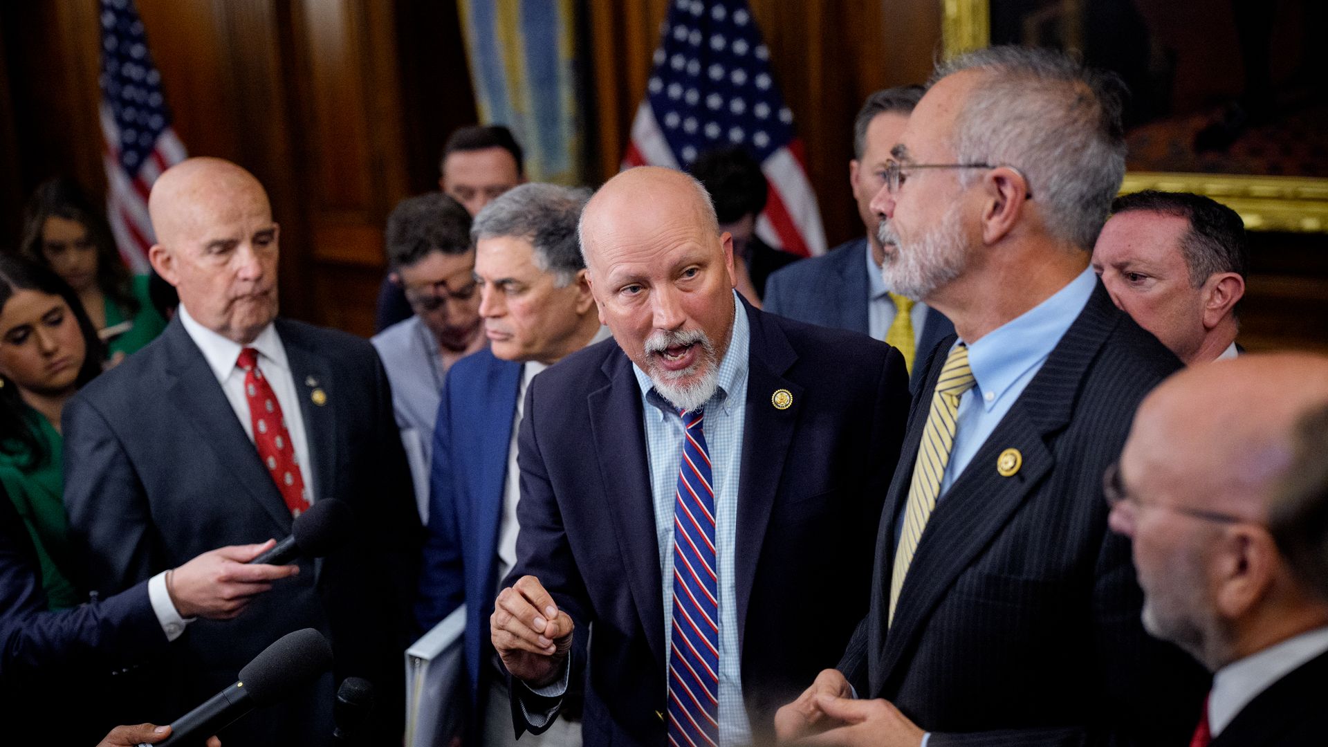 Group of men in suits and ties speaking to reporters with microphones in an indoor setting decorated with American flags and wood paneling.