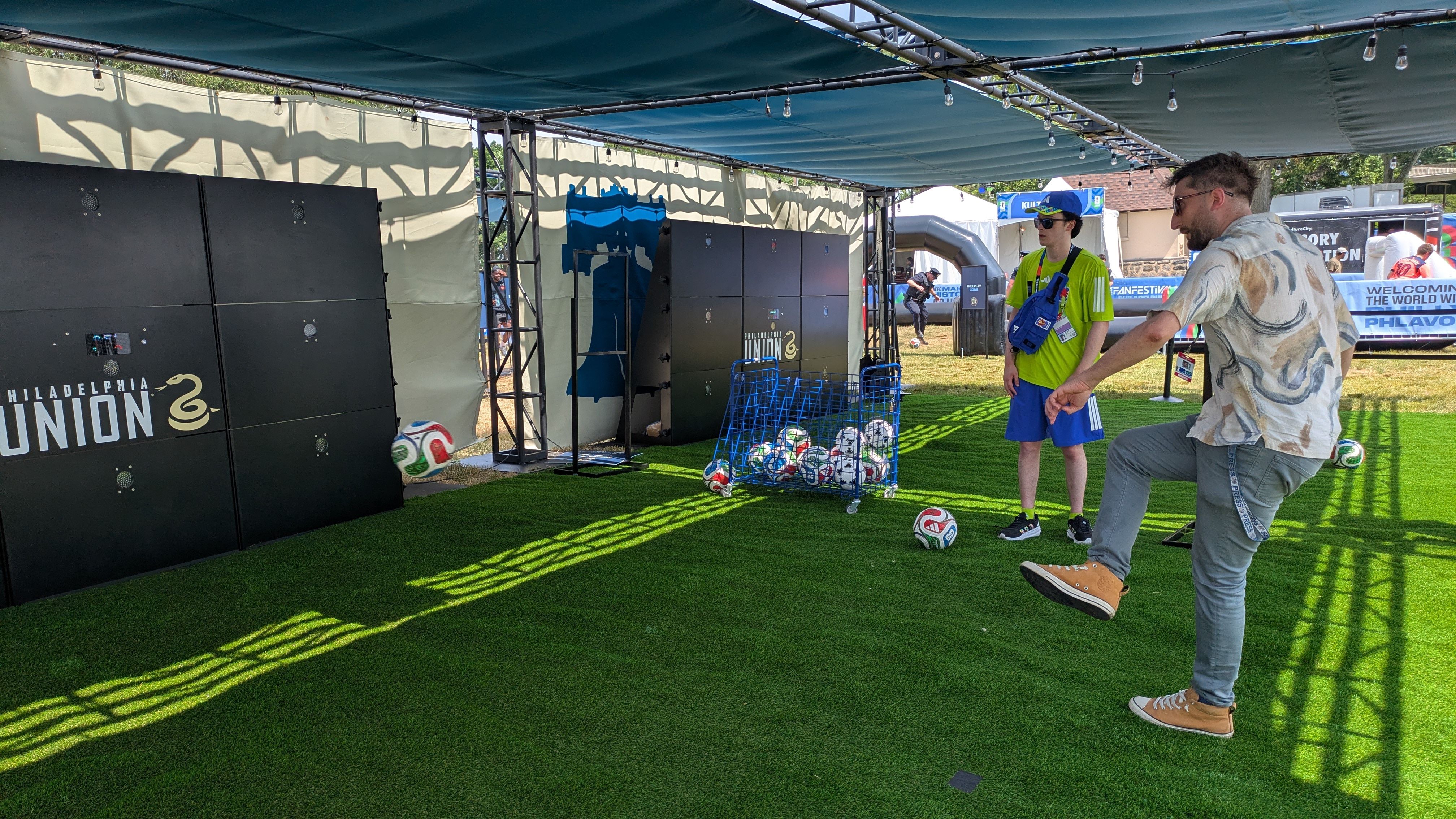 Outdoor event under a teal canopy with string lights. Large black Philadelphia Union lockers line the wall, next to a blue ball cart; two men watch as another kicks a soccer ball on green turf.