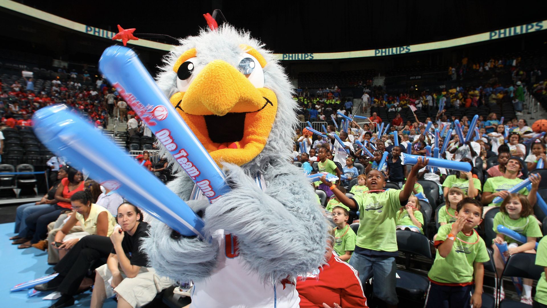 A bird mascot holding two sky blue inflatable noisemakers reading "One team" stands in front of a group of cheering children wearing neon green t-shirts and holding similar objects.