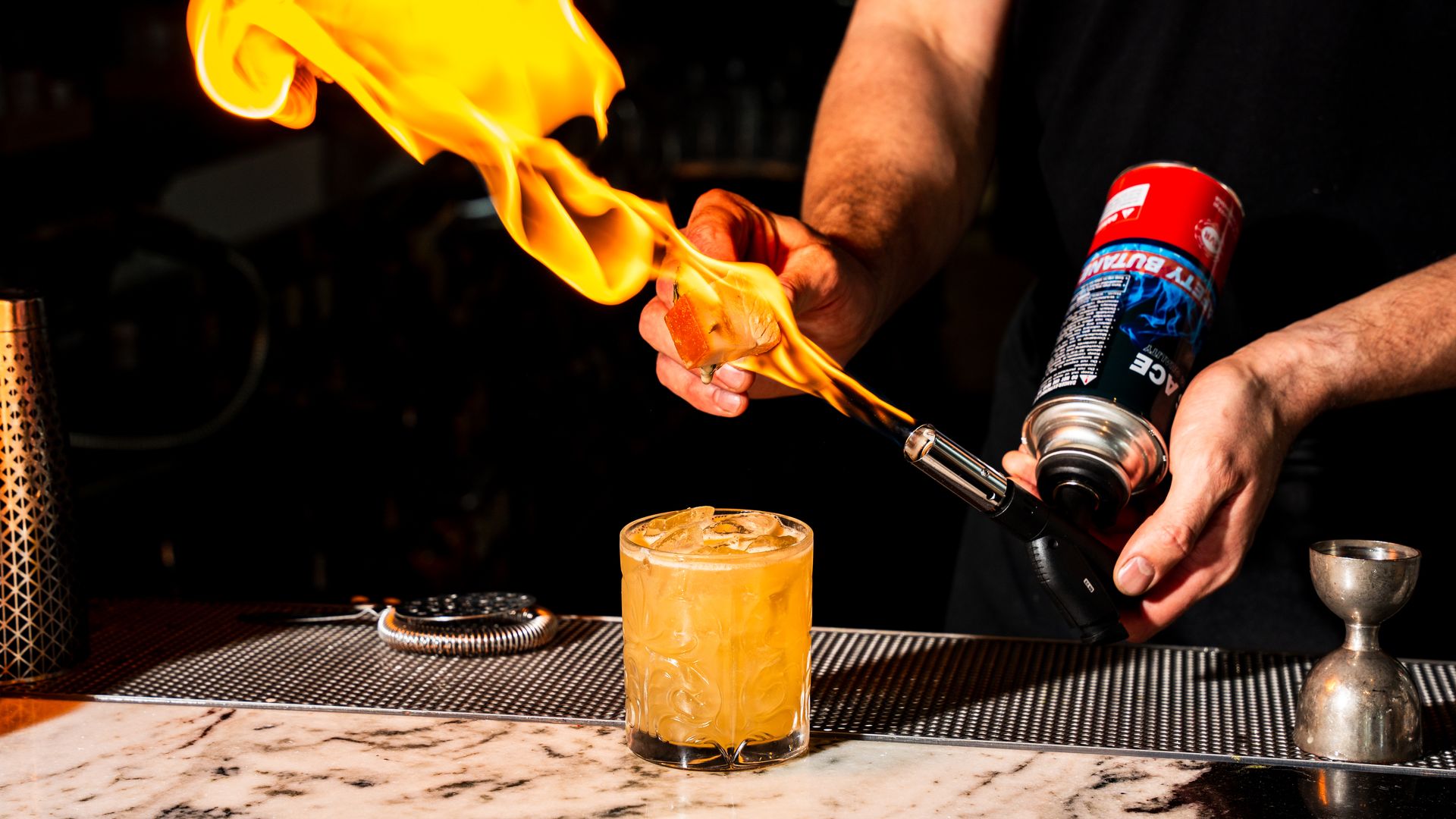 A bartender lights a garnish on fire above a yellow cocktail