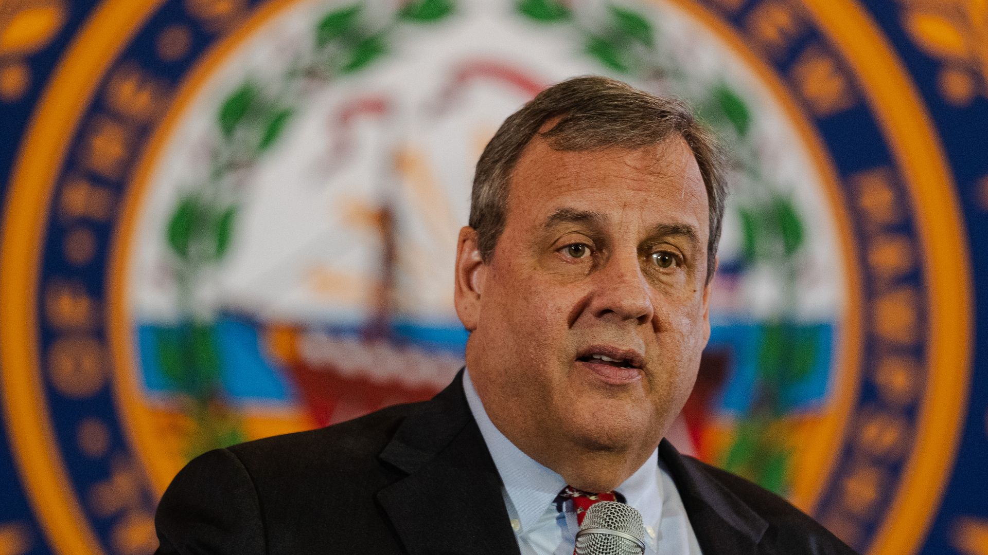 Man in dark suit and red tie holding a microphone, Chris Christie, former governor of New Jersey, speaking with a seal featuring green leaves and a ship in the blurred background.
