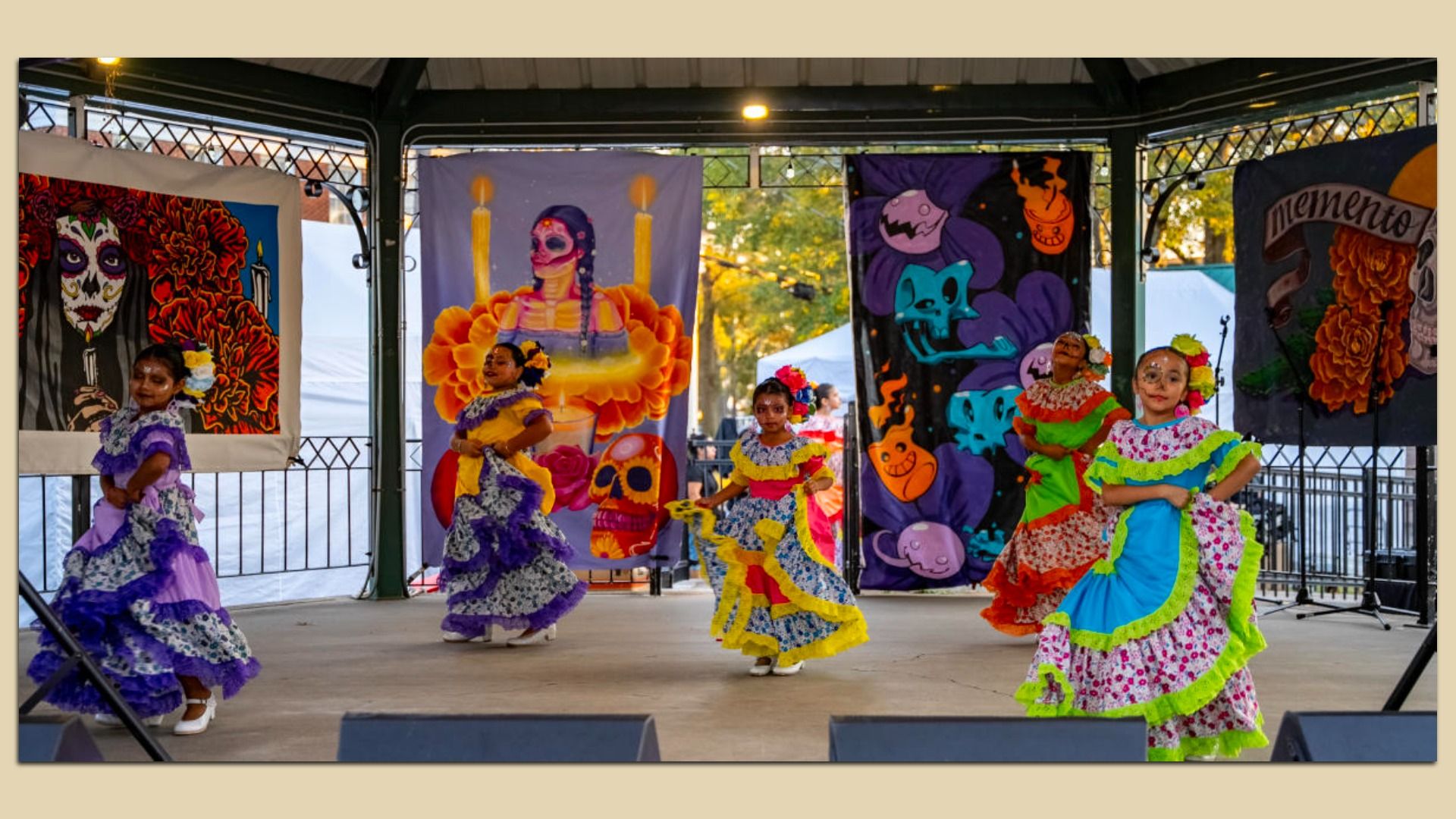 Young girls in colorful traditional dresses with face paint perform folkloric dance on a stage decorated with vibrant Día de los Muertos-themed banners featuring skulls and marigolds.