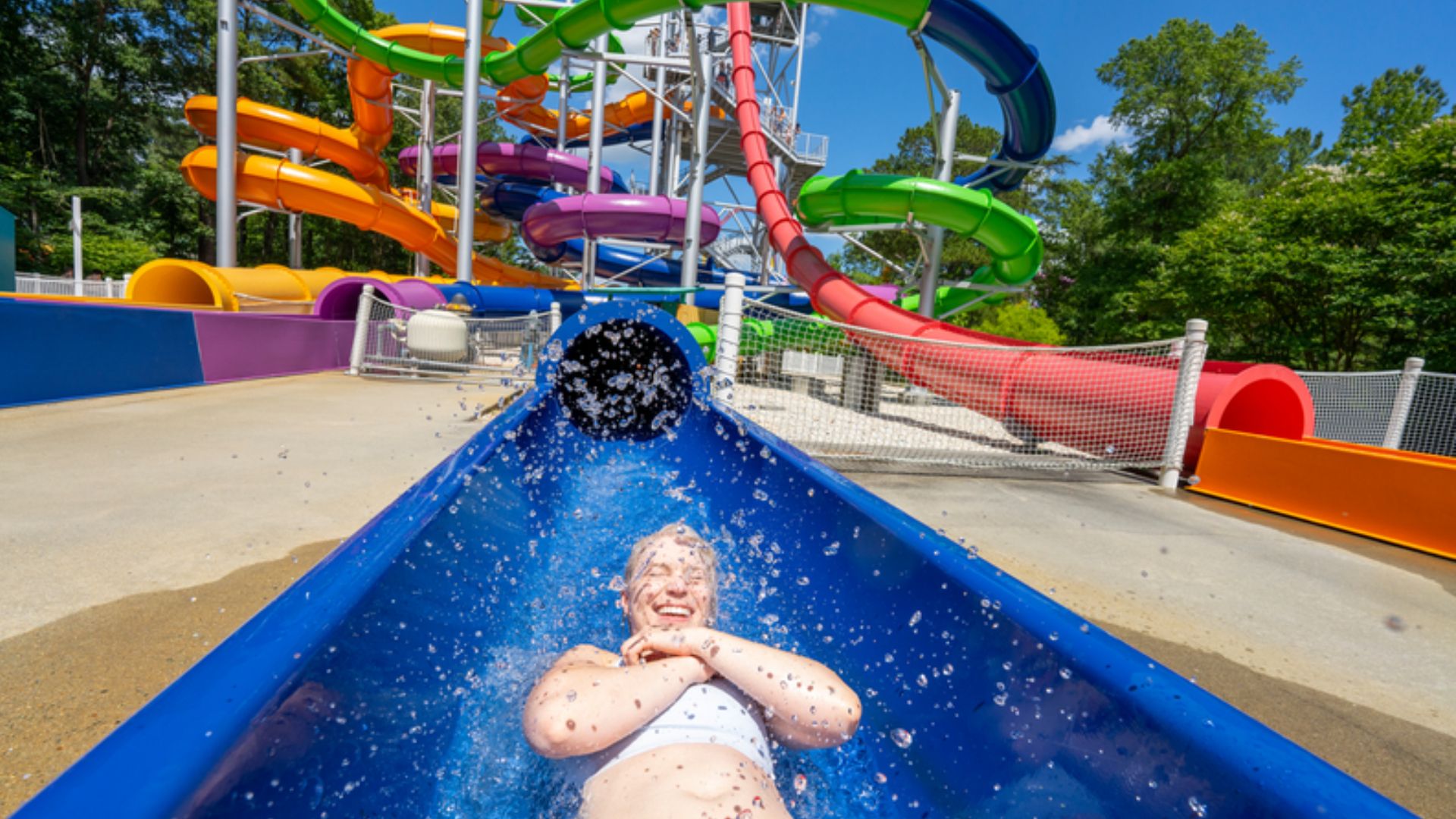 Person wearing a white swimsuit slides down a bright blue water slide at a sunny outdoor water park, with colorful twisting slides (orange, pink, green) and green trees in the background.