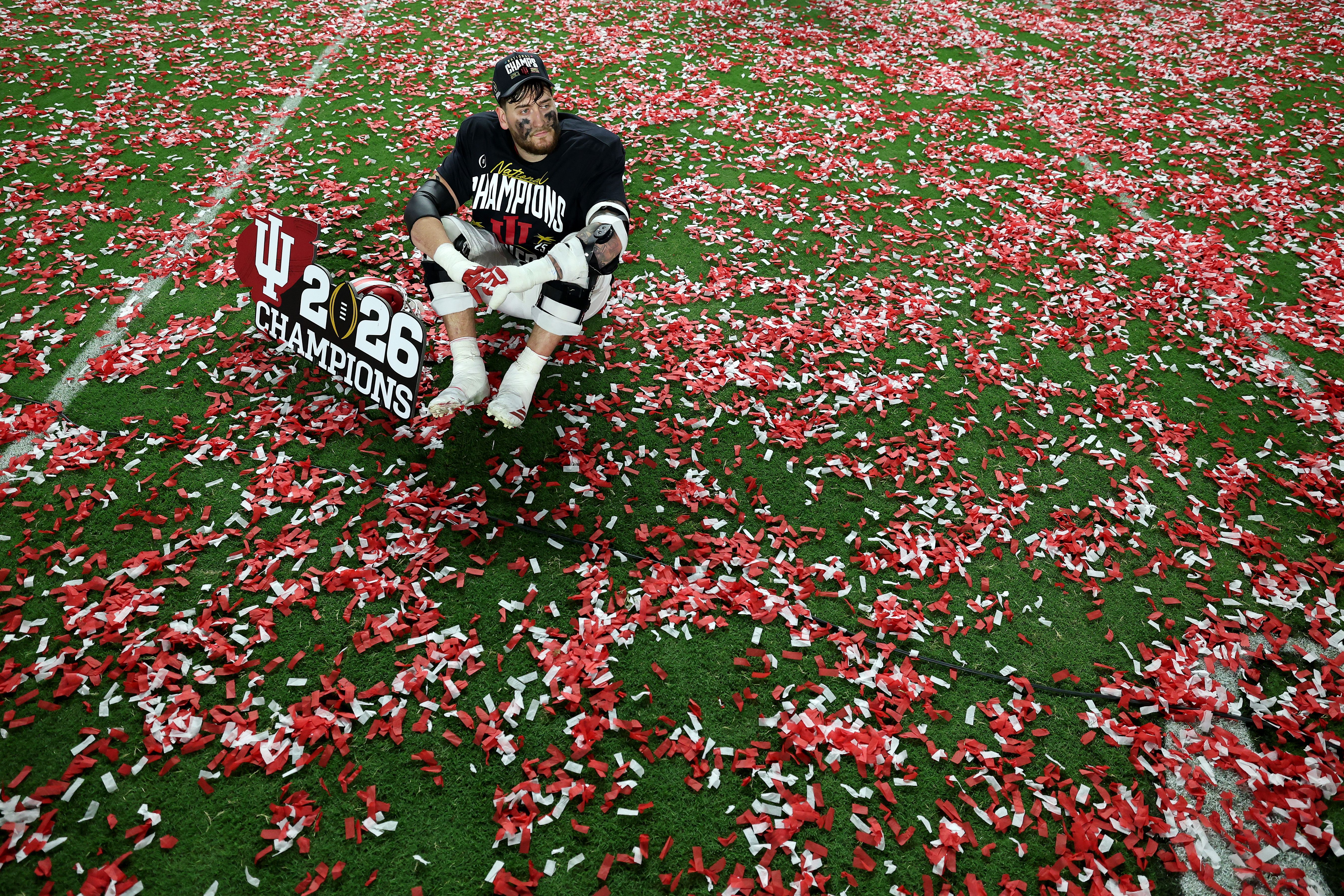Football player in black "National Champions" shirt and cap sitting on field covered in red and white confetti, holding a sign reading "IU 26 Champions".