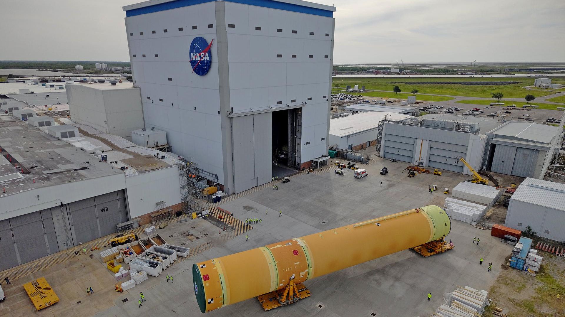 The large Artemis III core stage, a component of the space launch system, is shown being rolled out of a large square NASA building. 