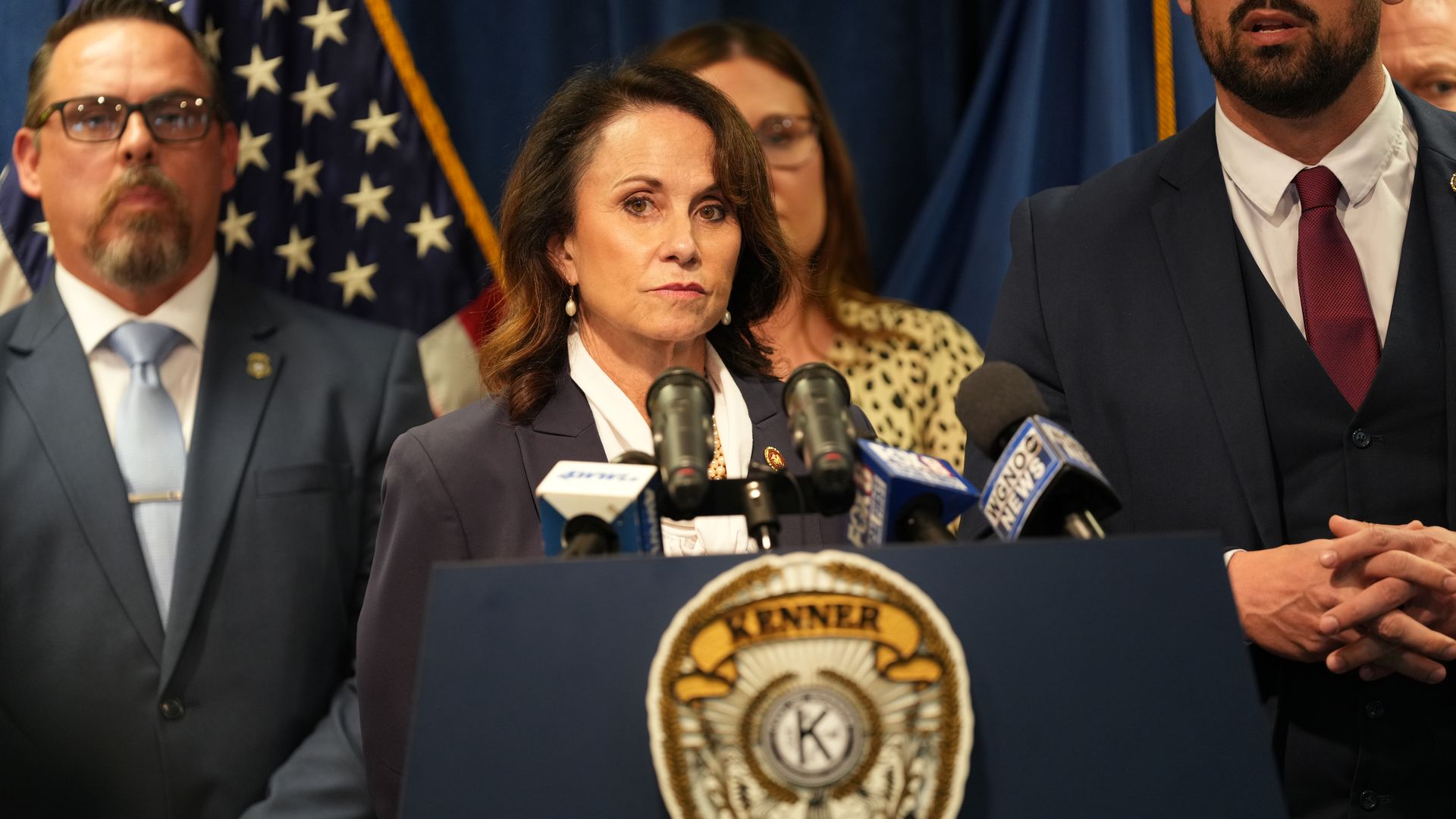 Centered woman in a dark blazer speaks at a police podium with a large crest, flanked by two men in suits. Microphones and American flags frame a formal press conference scene.