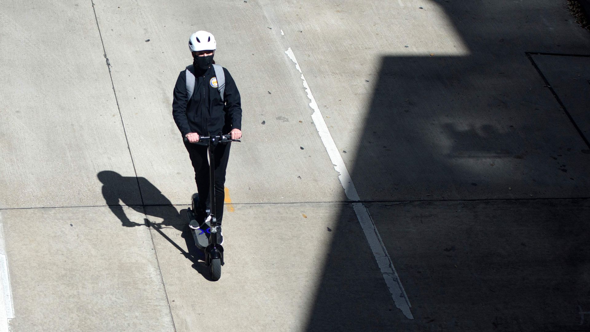 Person in black clothing and white helmet riding an electric scooter on sunlit, empty concrete street with large dark shadows cast on road.
