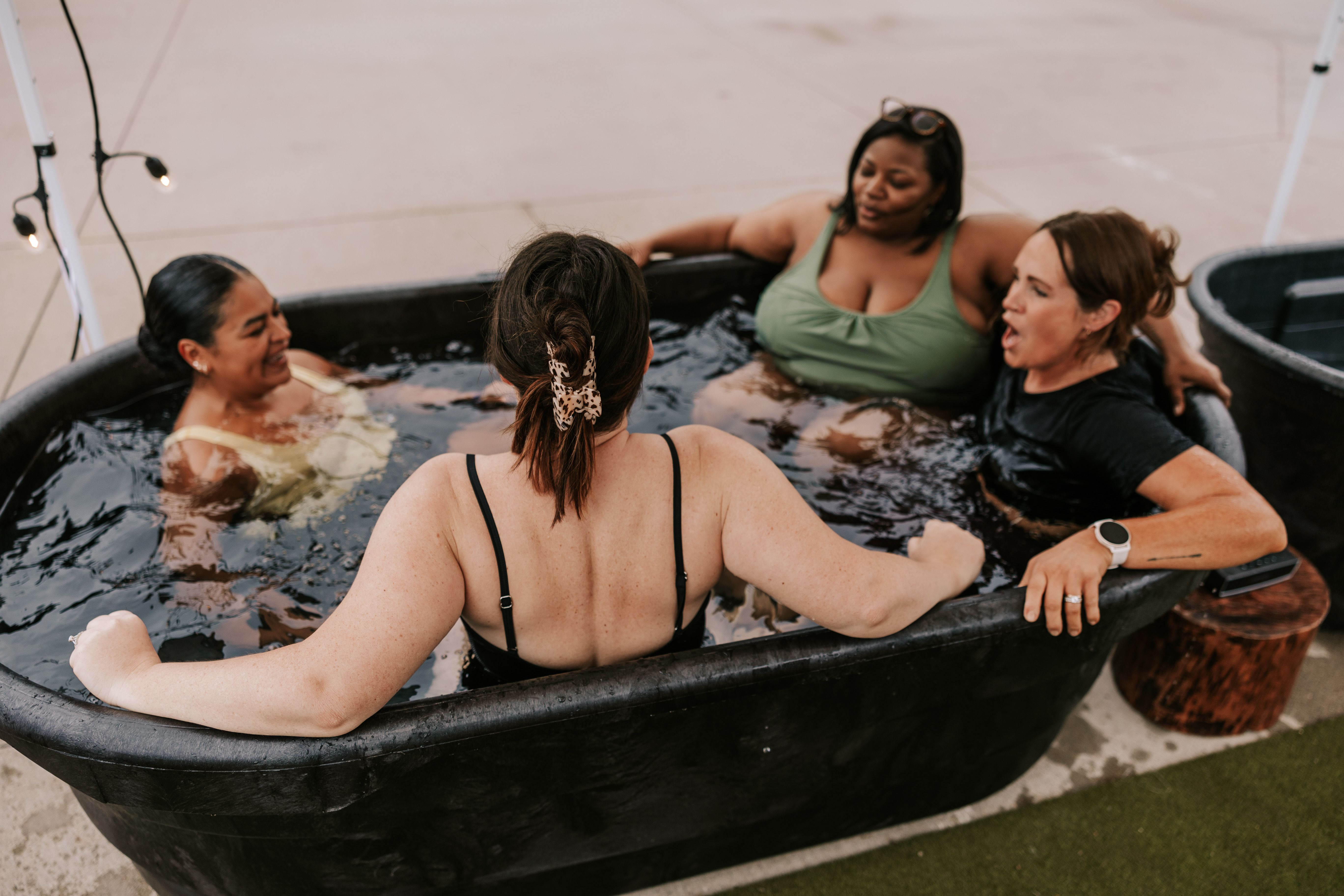 Four women sit in a black outdoor tub on a concrete patio, chatting and laughing. One with a leopard hair clip faces away; others wear swimsuits as string lights glow nearby.