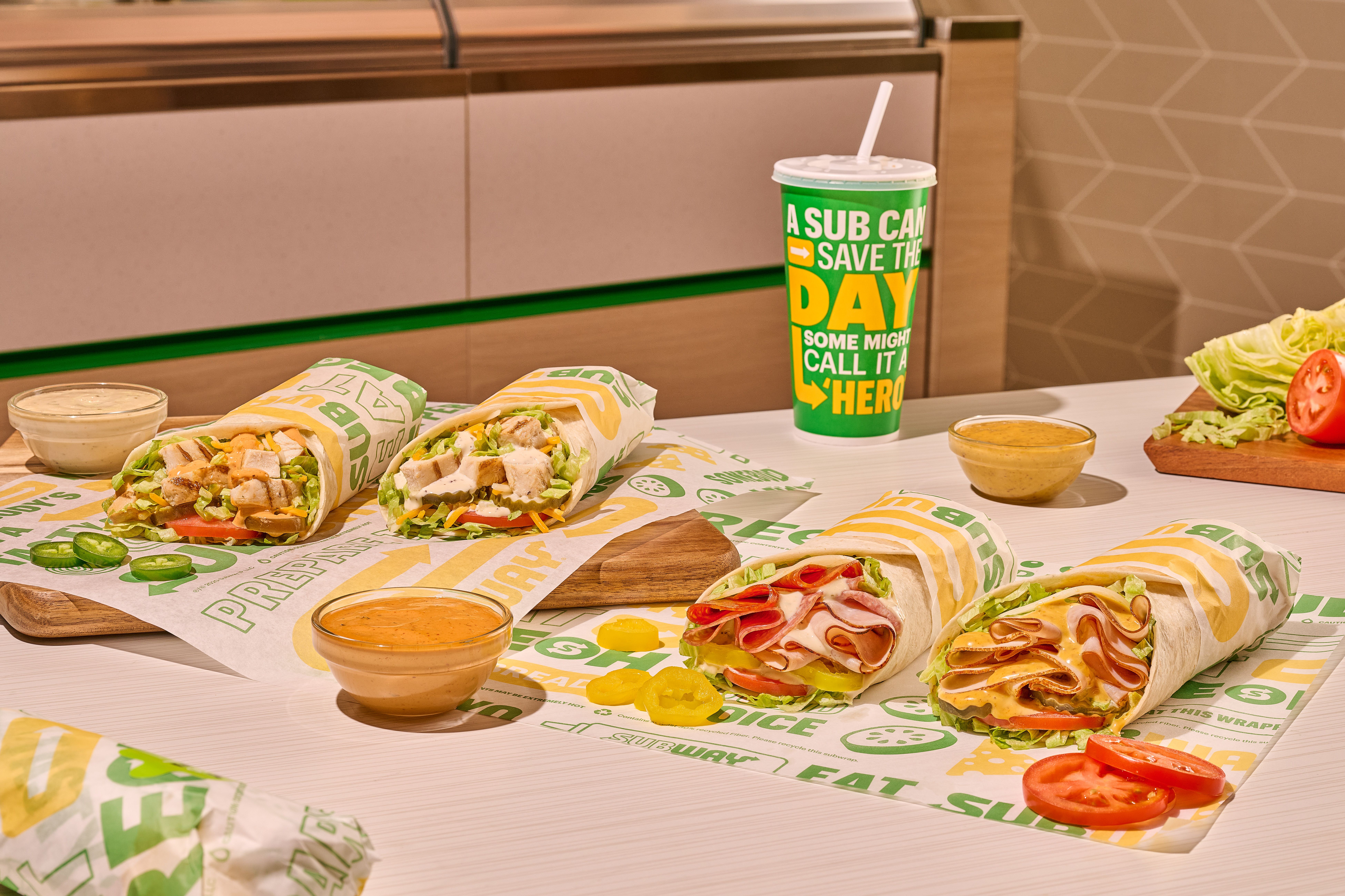Four Subway sandwiches on branded paper with dipping sauces, sliced jalapeños, tomatoes, lettuce, and a large green drink cup that reads "A SUB CAN SAVE THE DAY, SOME MIGHT CALL IT A HERO"