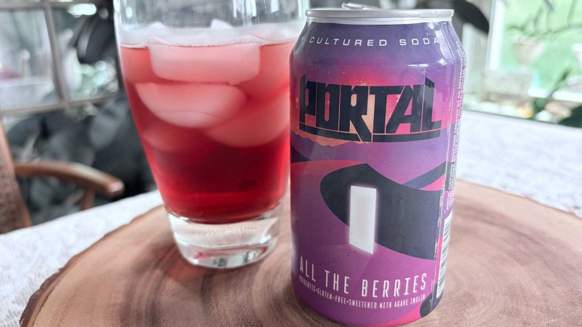 Purple can labeled "All The Berries" Cultured Soda beside a glass of pink drink with ice on a wooden table; blurred plants and windows in the background.