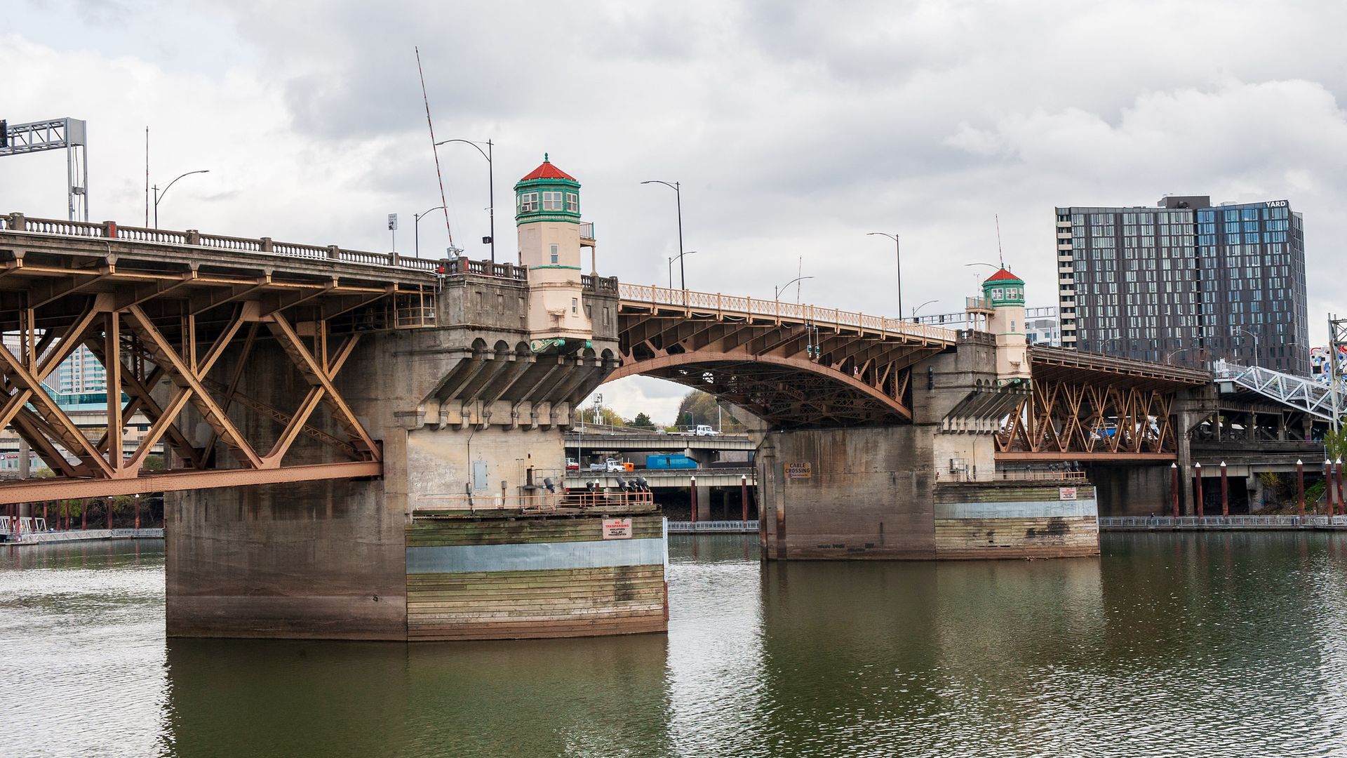 Brown steel bridge over a calm river, with beige towers featuring green trim and red roofs; a modern glass building skyline rises in the cloudy city backdrop.
