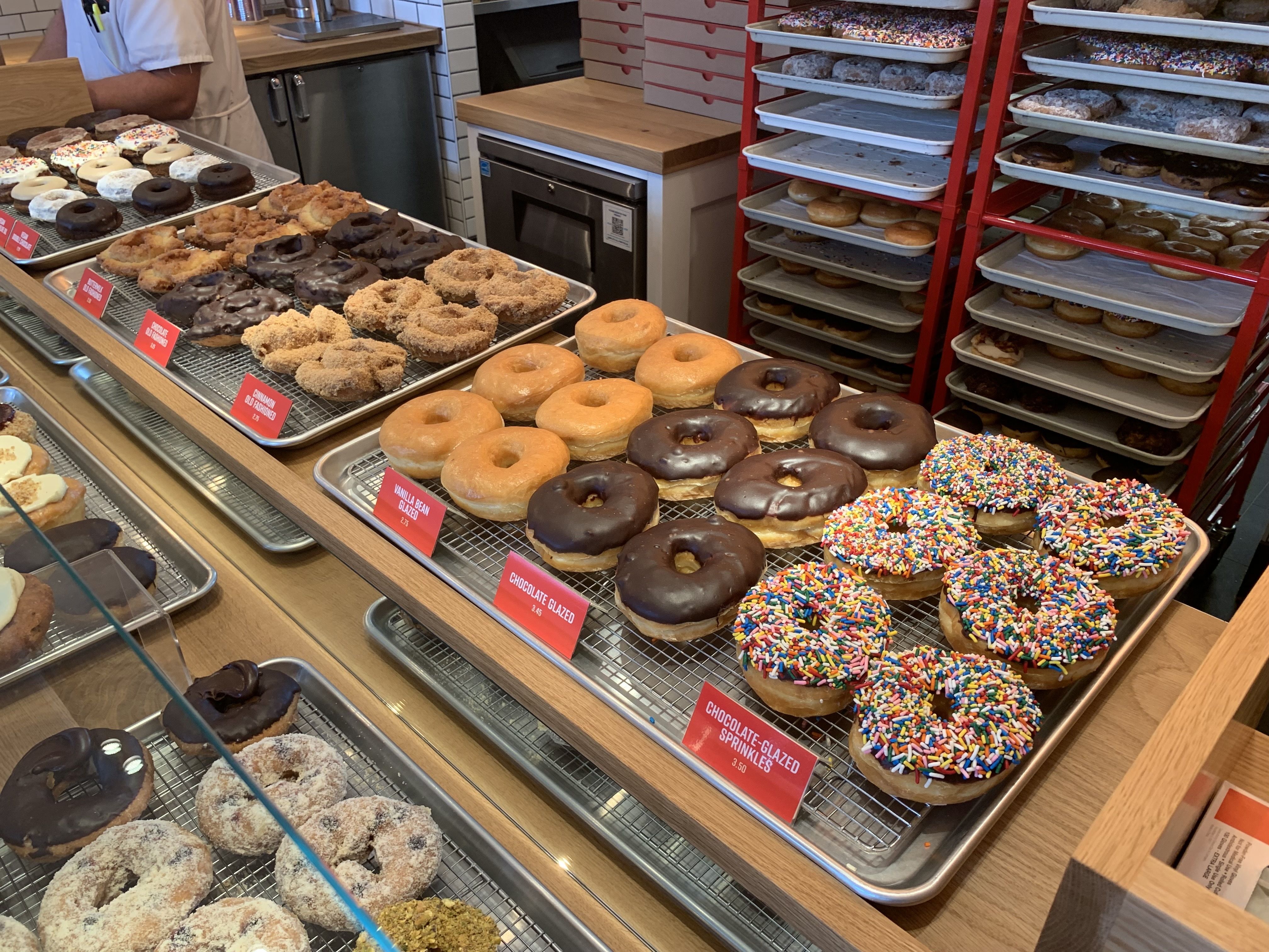 Display of assorted doughnuts on trays at a bakery counter, including chocolate glazed, vanilla bean glazed, plain glazed, and rainbow-sprinkled varieties; red racks full of donuts in the background.