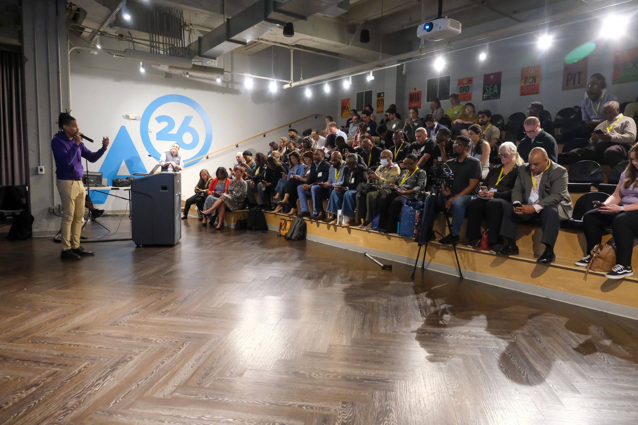 Speaker in a purple hoodie at a podium addresses a crowded audience in a tiered lecture hall; a large blue graphic with "A26" and travel banners adorn the wall.