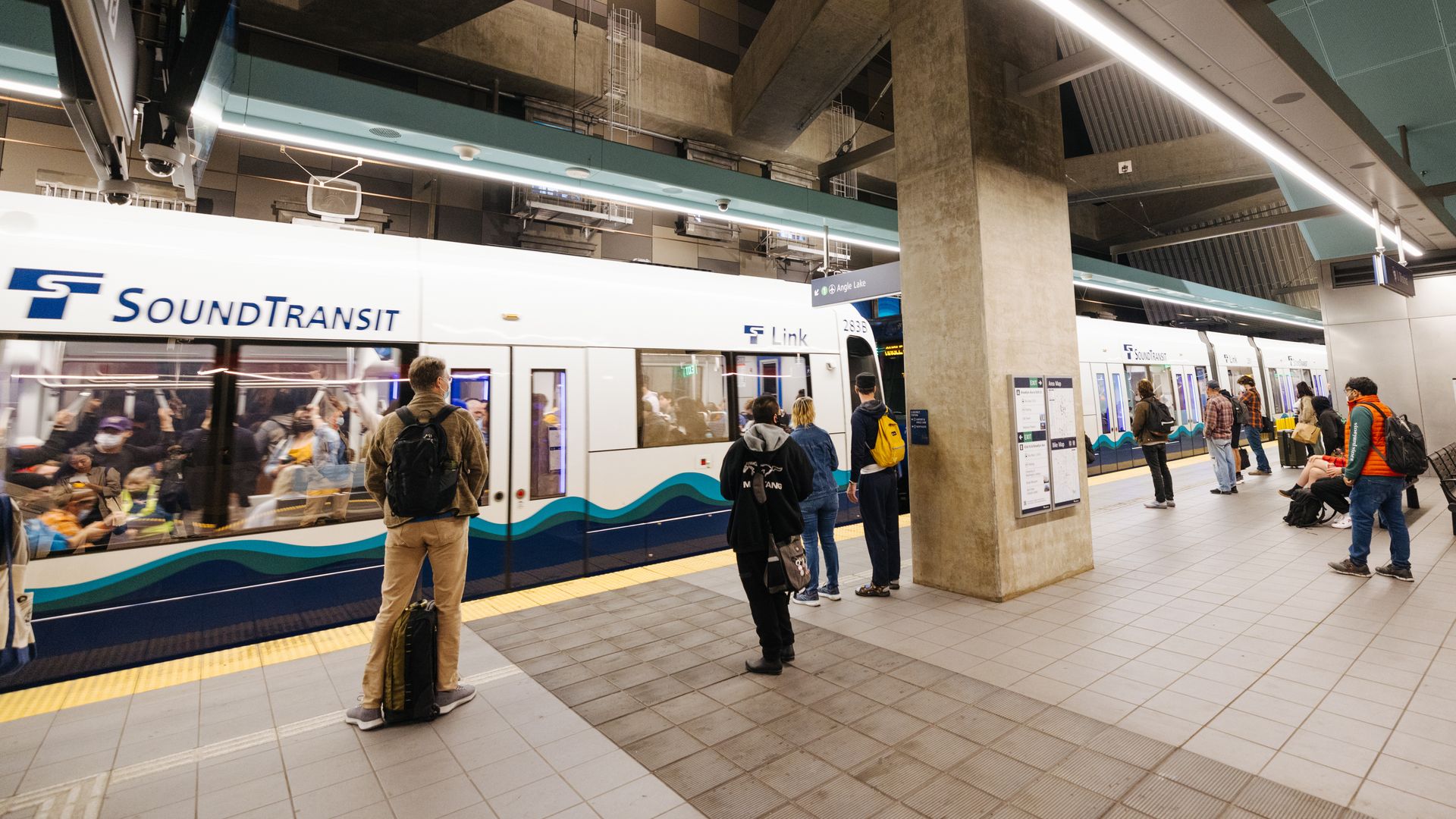 People wait on a tiled platform at an underground Sound Transit station, near a white and blue Link light rail train with windows showing seated passengers inside.