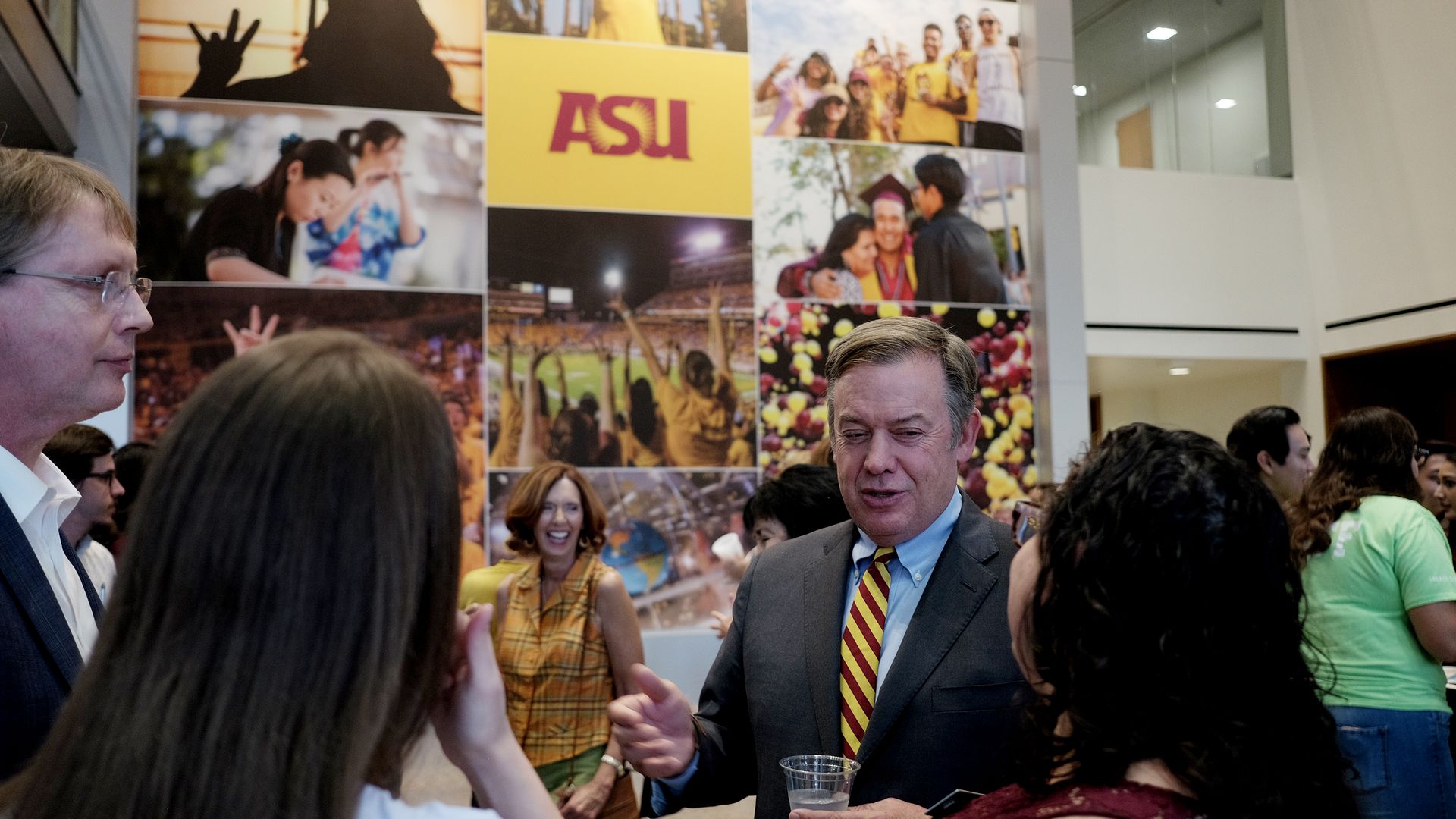 A man in a suit speaks to several people in front of a large sign that reads A S U.