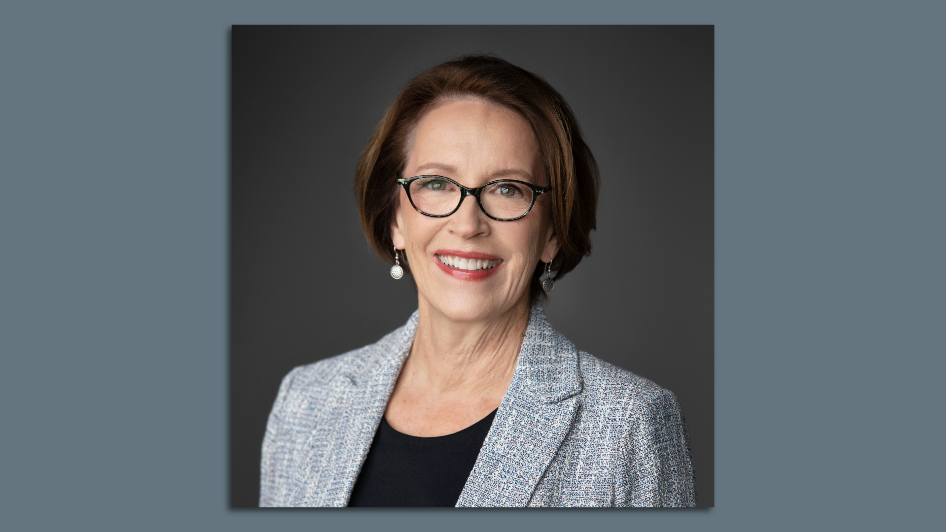 Smiling middle-aged woman with short brown hair, black-rimmed glasses, and a light blue tweed blazer over a black top, set against a dark gray gradient background.