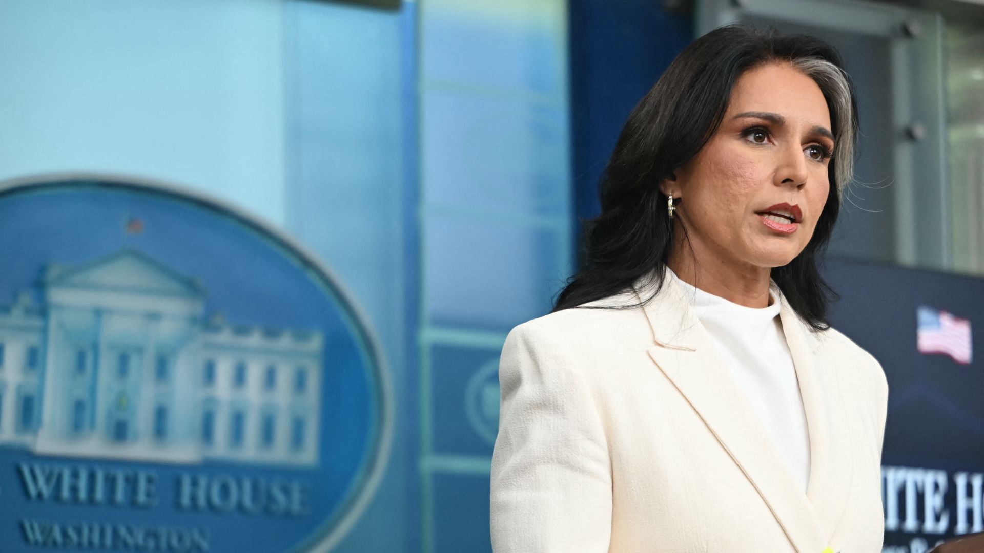 Woman in a white blazer speaking at a White House podium, with the White House emblem in blue visible on the wall behind her.