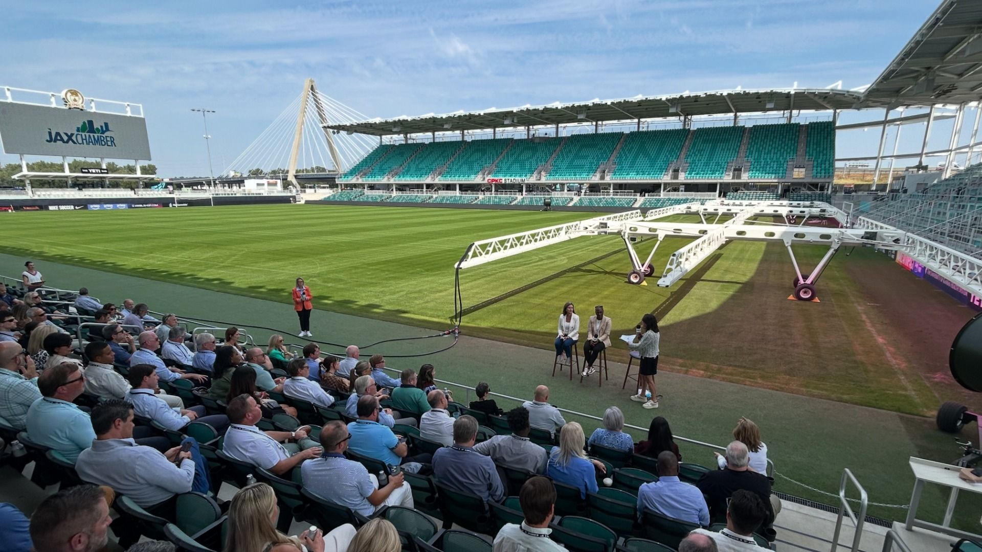 Bright stadium with green grass and teal seating. A long white maintenance arm spans the field while four people sit on stools for a panel; a crowd watches from the stands and a "JAX Chamber" sign is visible.