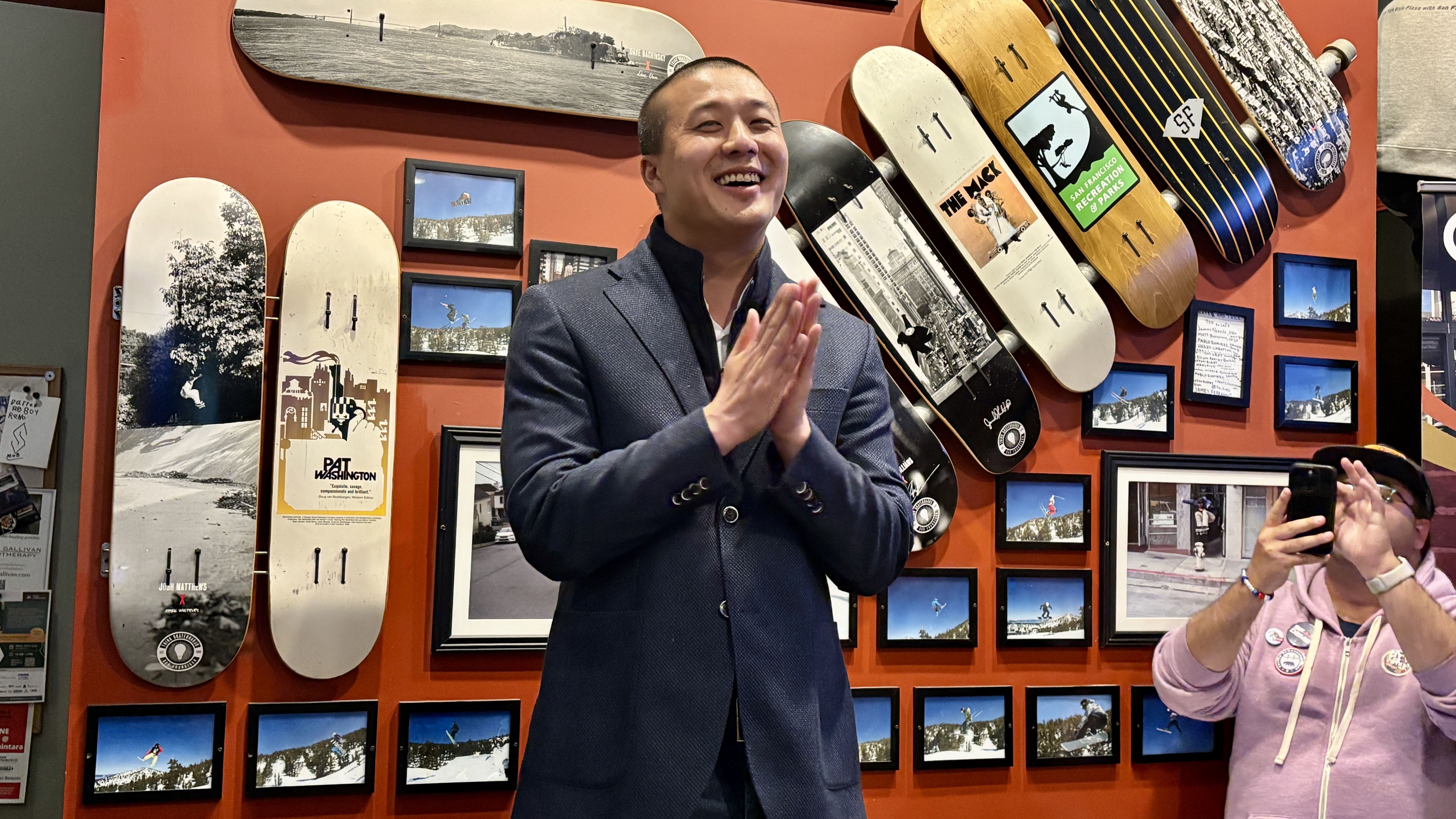 Smiling man in a blue blazer stands with hands pressed together in front of a red wall covered with skateboard decks and framed photos; another person in pink holds a phone.