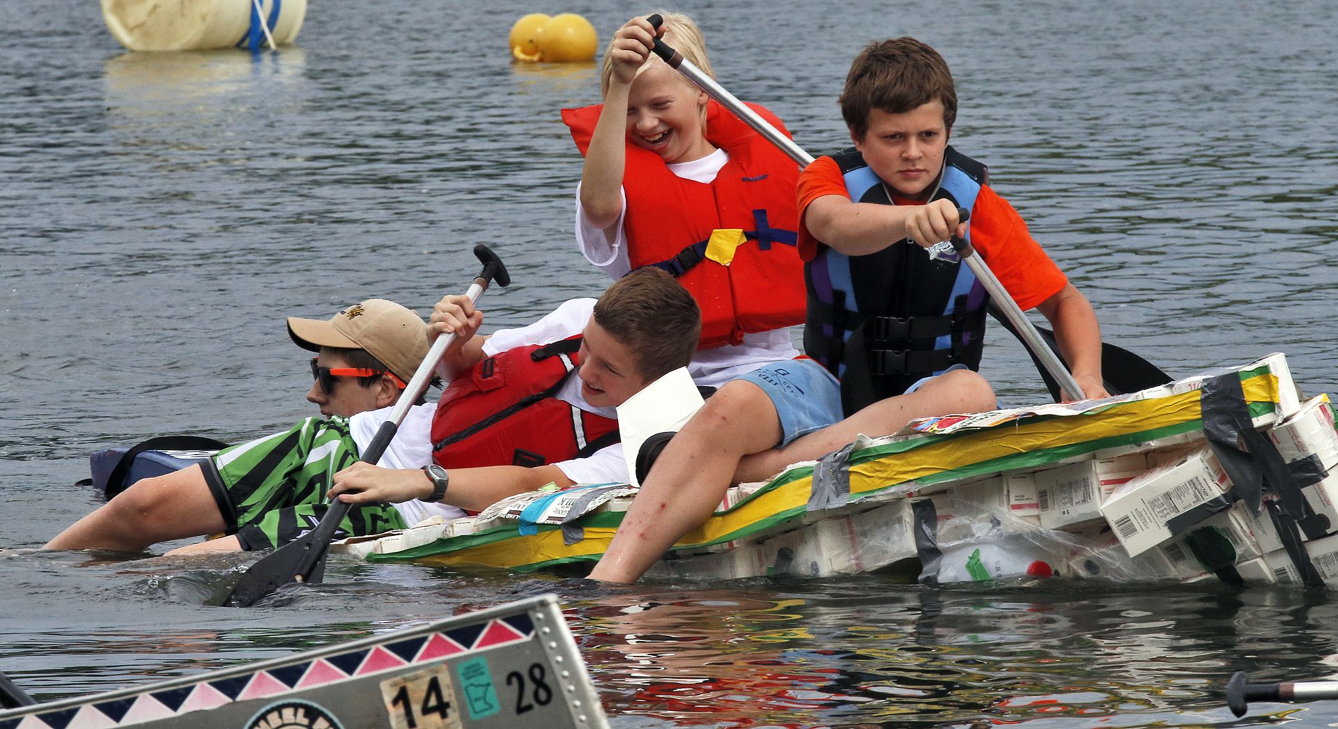 Four kids in bright life jackets paddle a homemade milk carton raft taped with colorful strips on a lake, racing with oars beside floating yellow buoys.