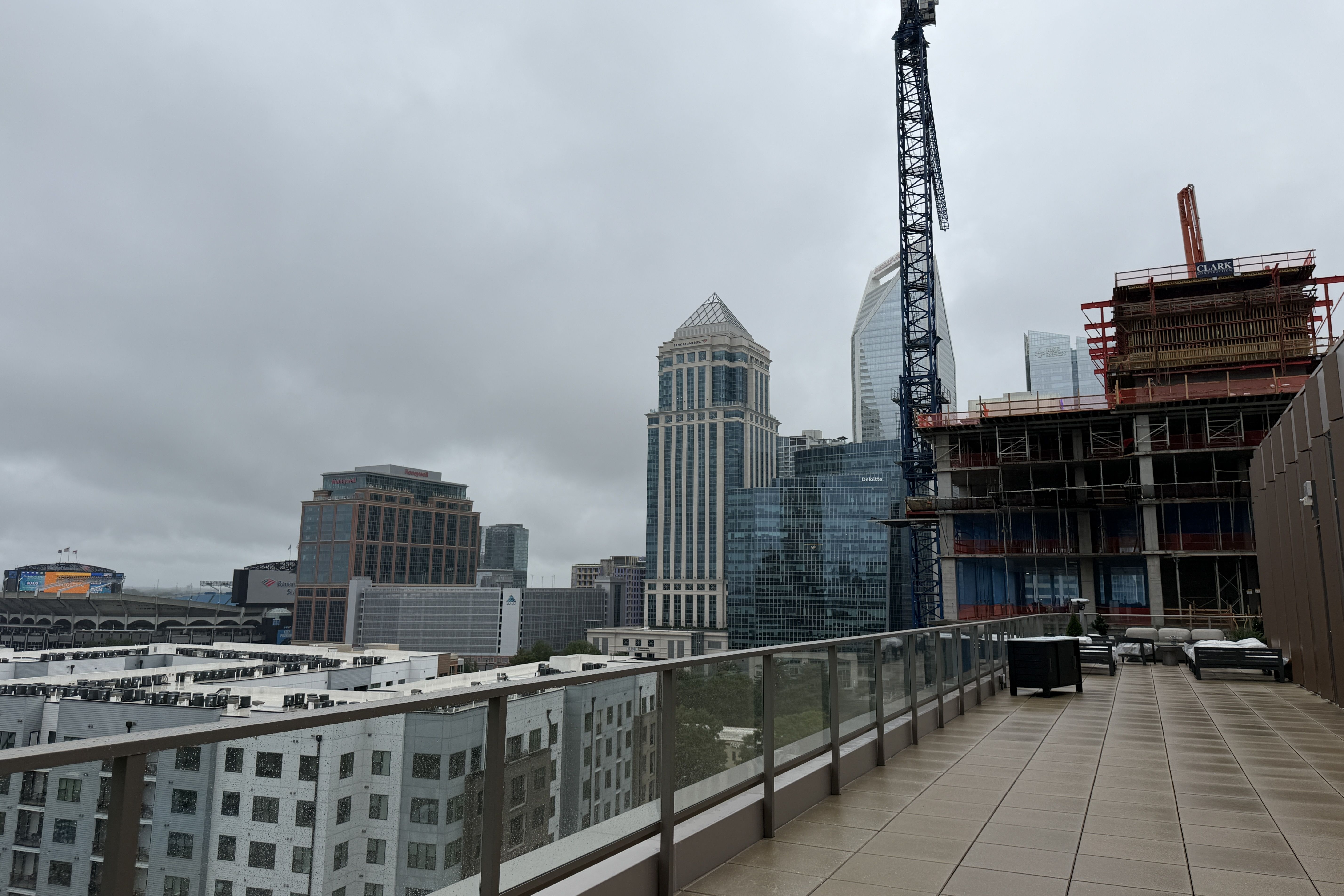 Gray, cloudy city skyline with a tall construction crane and orange scaffolding on the right; glass skyscrapers rise in the distance, and a rooftop terrace with glass railing sits in the foreground.