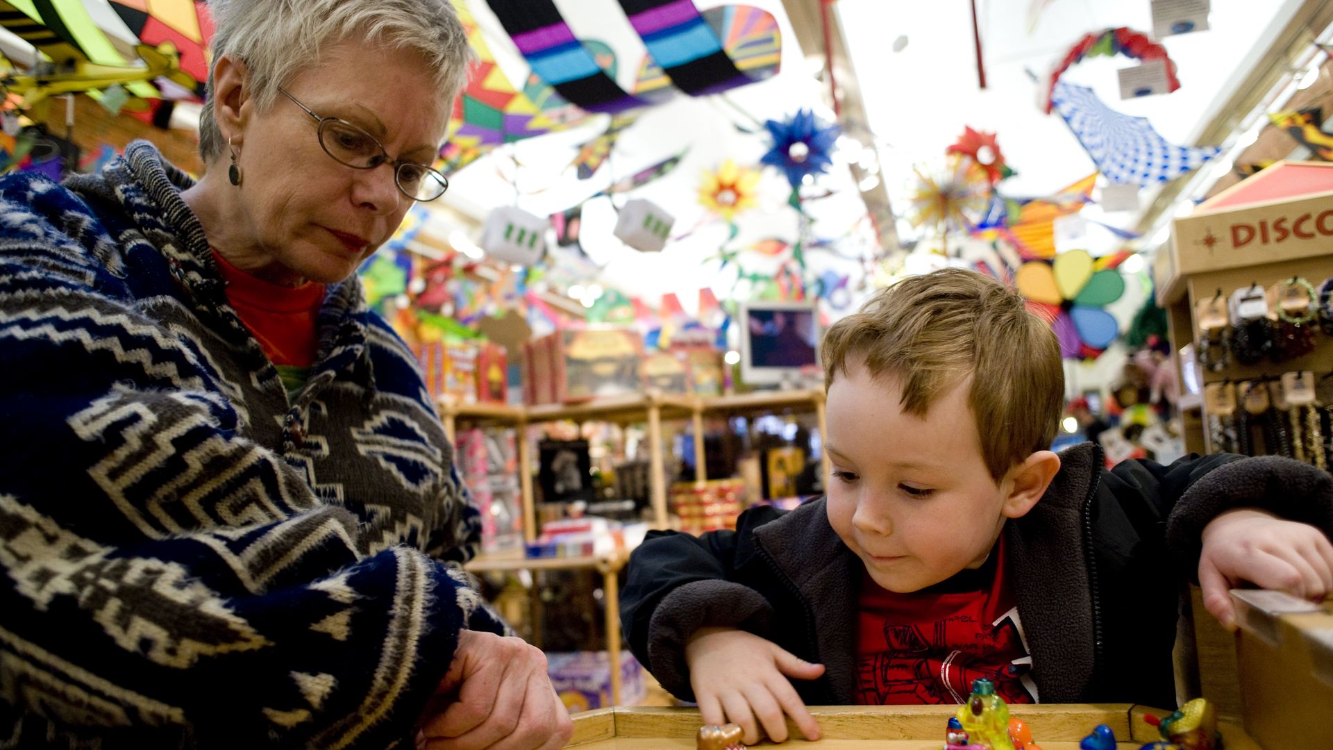 A woman and a young boy looking intently at small colorful toys on a wooden display table inside a vibrant, decorated toy store with kites and various items hanging from the ceiling.