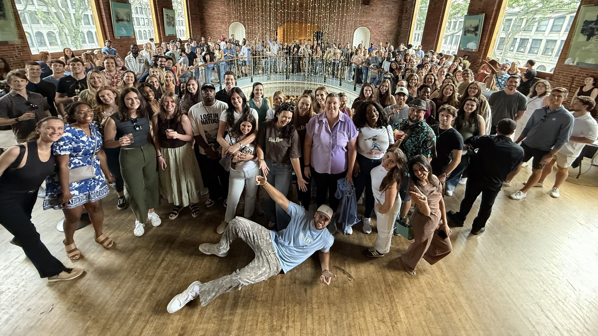 Large diverse group of people posing inside a brick building with tall arched windows and string lights hanging from the ceiling, smiling and looking at the camera.