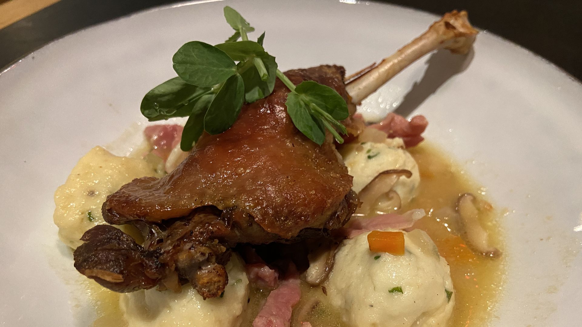 Plate with roasted duck leg garnished with greens, served on three fluffy dumplings in broth with sliced mushrooms, diced carrots, and bacon bits.