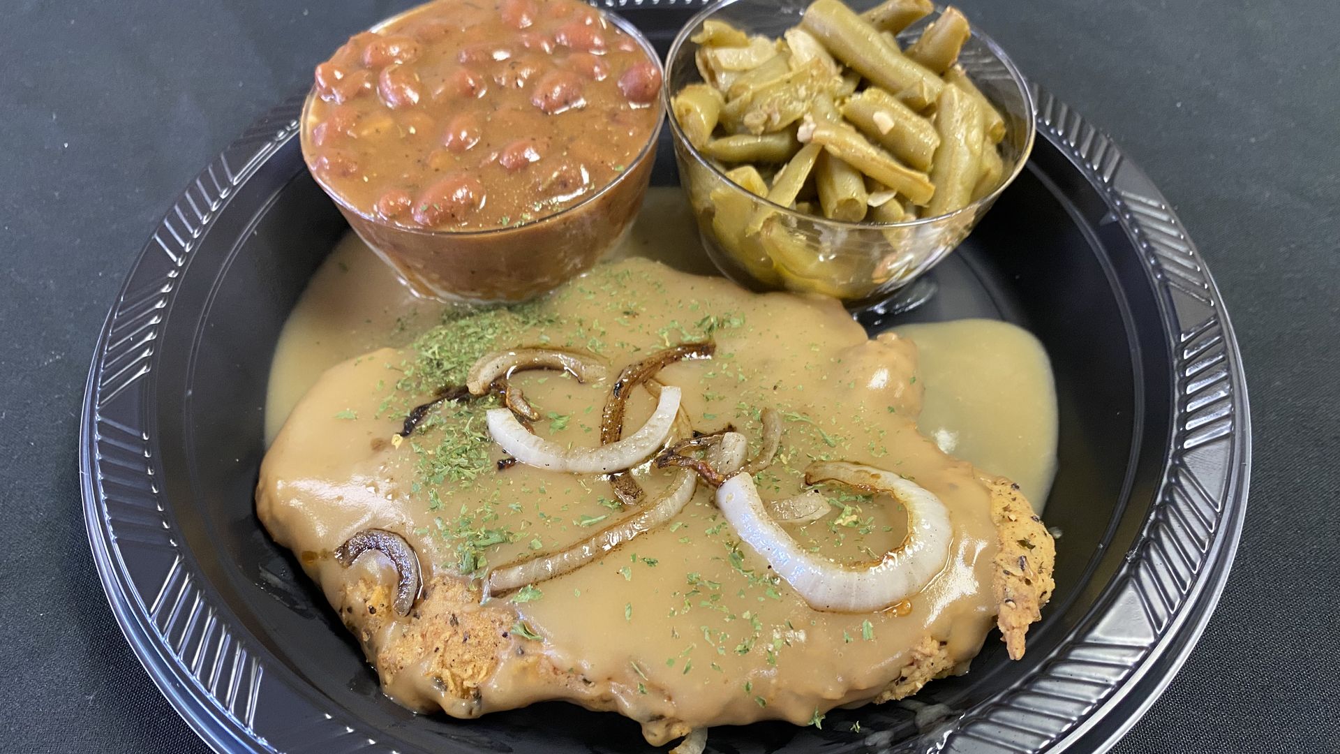 A piece of fried meat covered in brown gravy and onions, with cups of green beans and red beans.